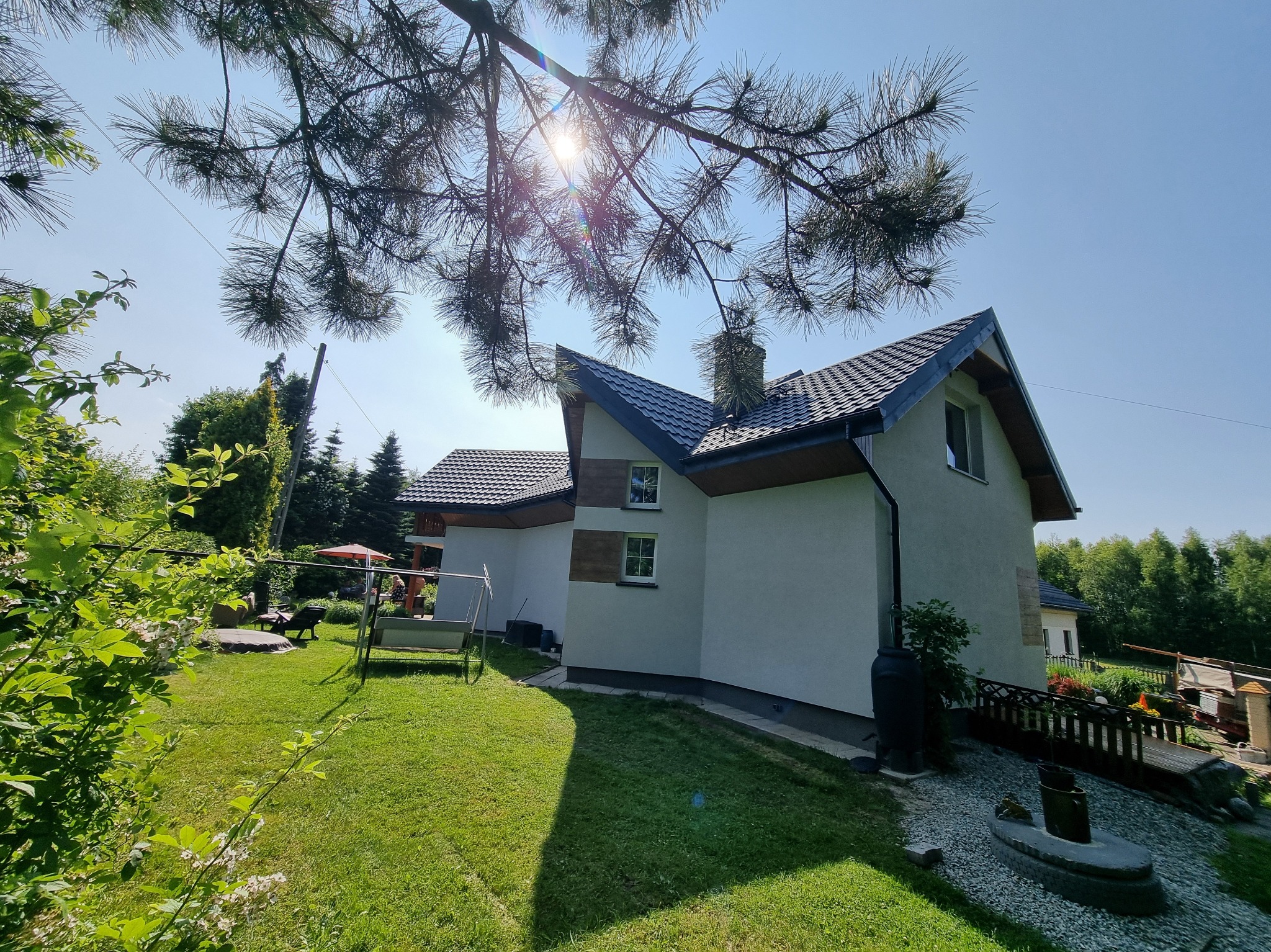 Dom jednorodzinny z ciemną dachówką w Wodzisławiu Śląskim, widziany z ogrodu przez gałęzie drzewa. Jasna elewacja, ciemne rynny i komin.