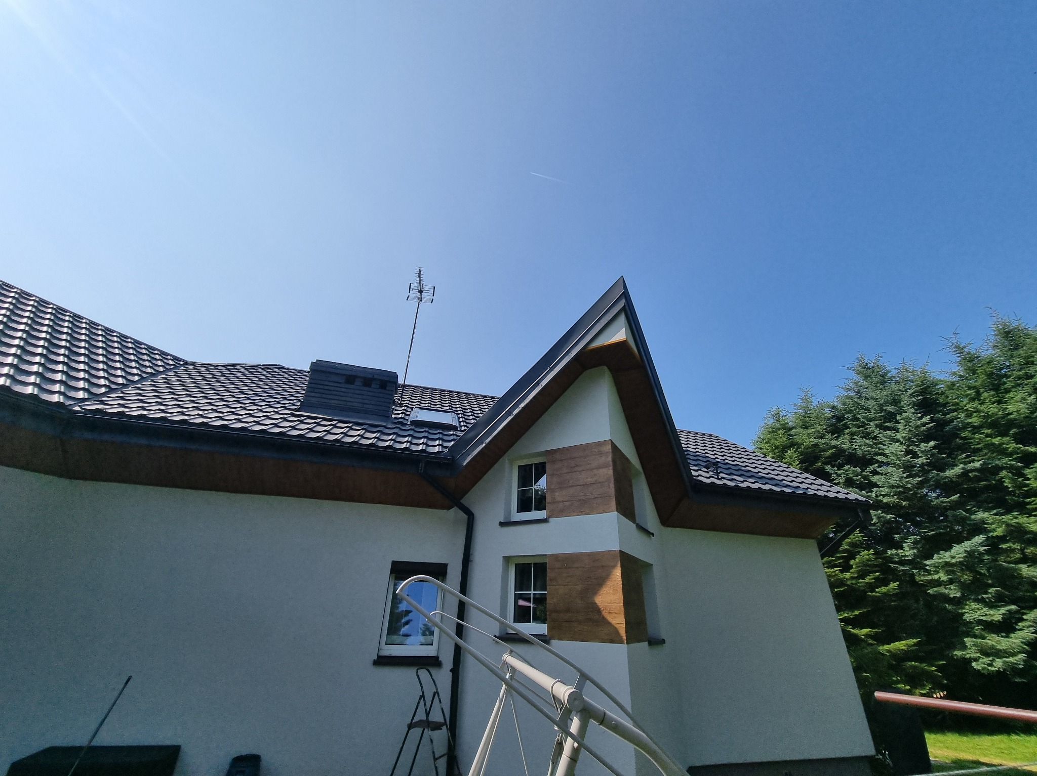 Dom z ciemną dachówką i drewnopodobną elewacją, widziany z dołu pod kątem. Na dachu antena, w tle błękitne niebo i zieleń drzew. Widoczne drabiny.