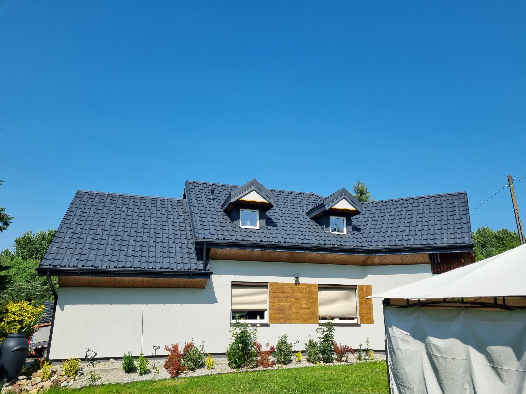 Dom jednorodzinny z ciemnoszarym dachem z dachówki ceramicznej, dwoma oknami dachowymi i jasną elewacją w słoneczny dzień. Widoczne obróbki blacharskie i rynny.
