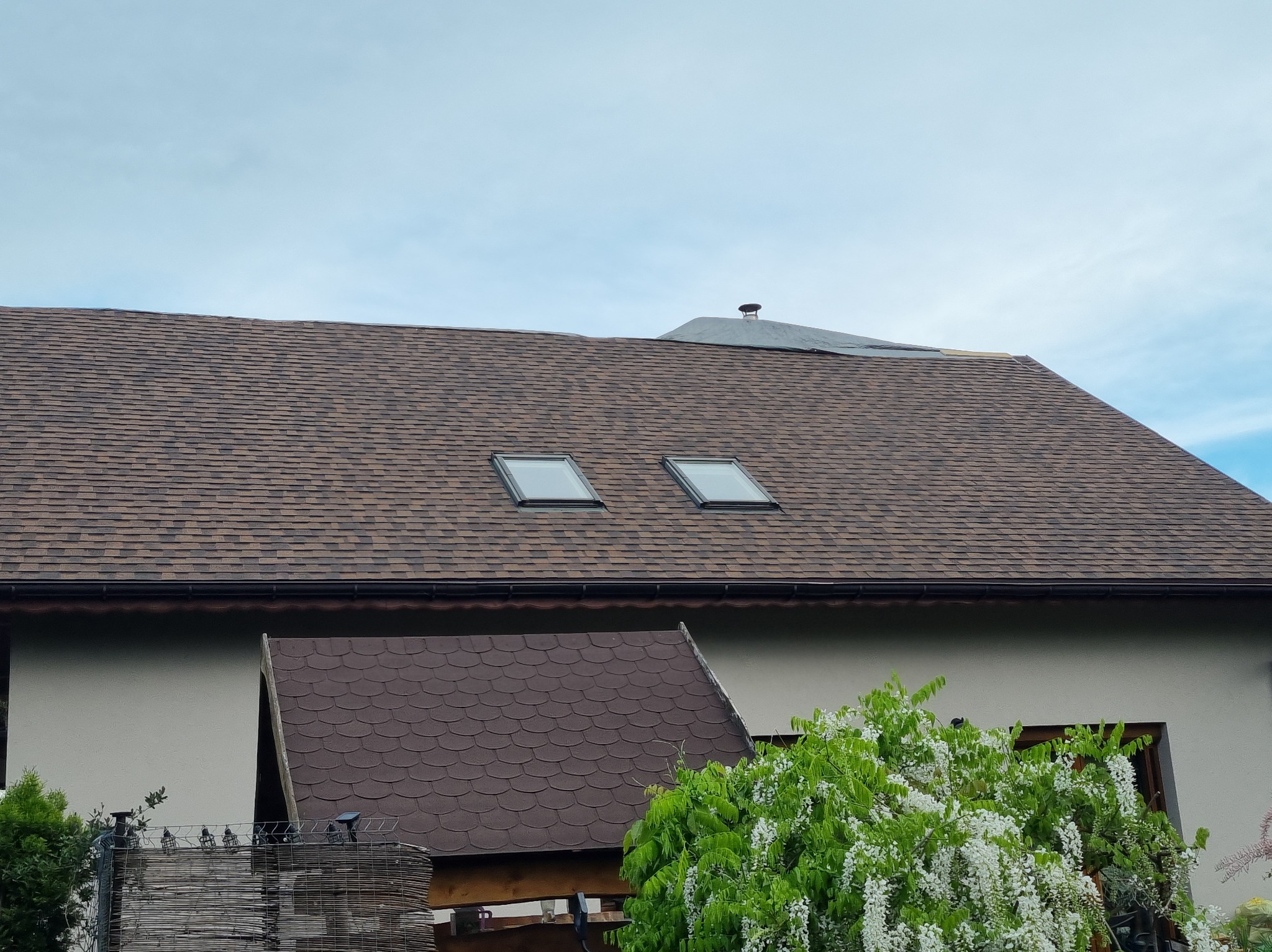 Dach pokryty brązowym gontem bitumicznym z dwoma oknami dachowymi. Fragment dachu w trakcie prac wykończeniowych. Widoczny komin i fragment elewacji budynku.