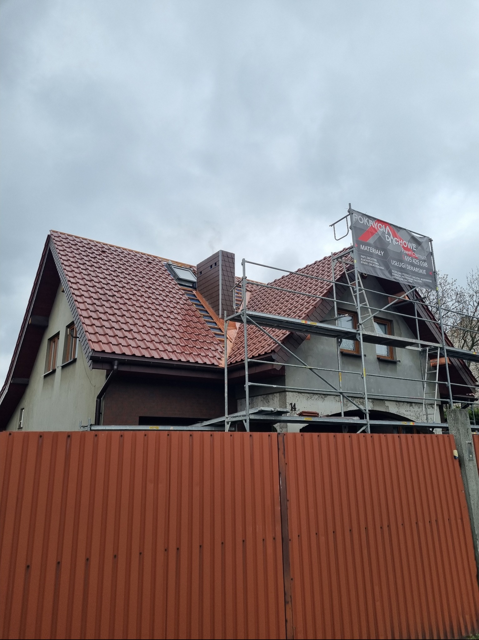 Dom z nowym, czerwonym dachem z dachówki, widoczny zza pomarańczowego ogrodzenia. Na dachu rusztowanie z banerem firmy dekarskiej.