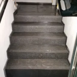 Schody wyłożone ciemnymi, teksturowanymi płytkami, widok z góry, z fragmentem narzędzia i pustaka budowlanego w tle, na ścianach widoczne ślady po taśmie malarskiej.