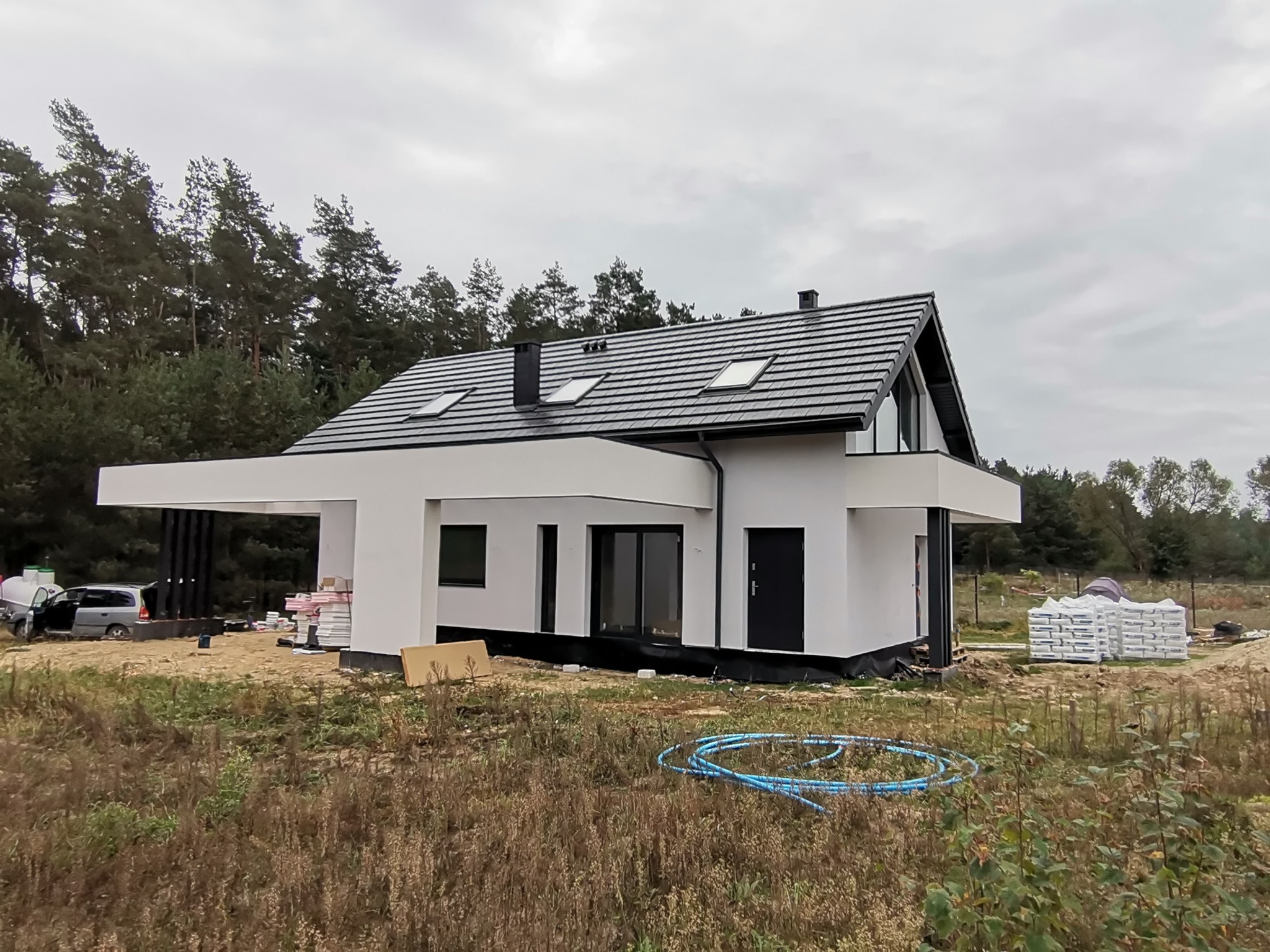 Nowoczesny, biały dom jednorodzinny z czarnym dachem i oknami dachowymi, częściowo ocieplony, z widocznymi materiałami budowlanymi na działce i niebieskimi rurami na pierwszym planie.