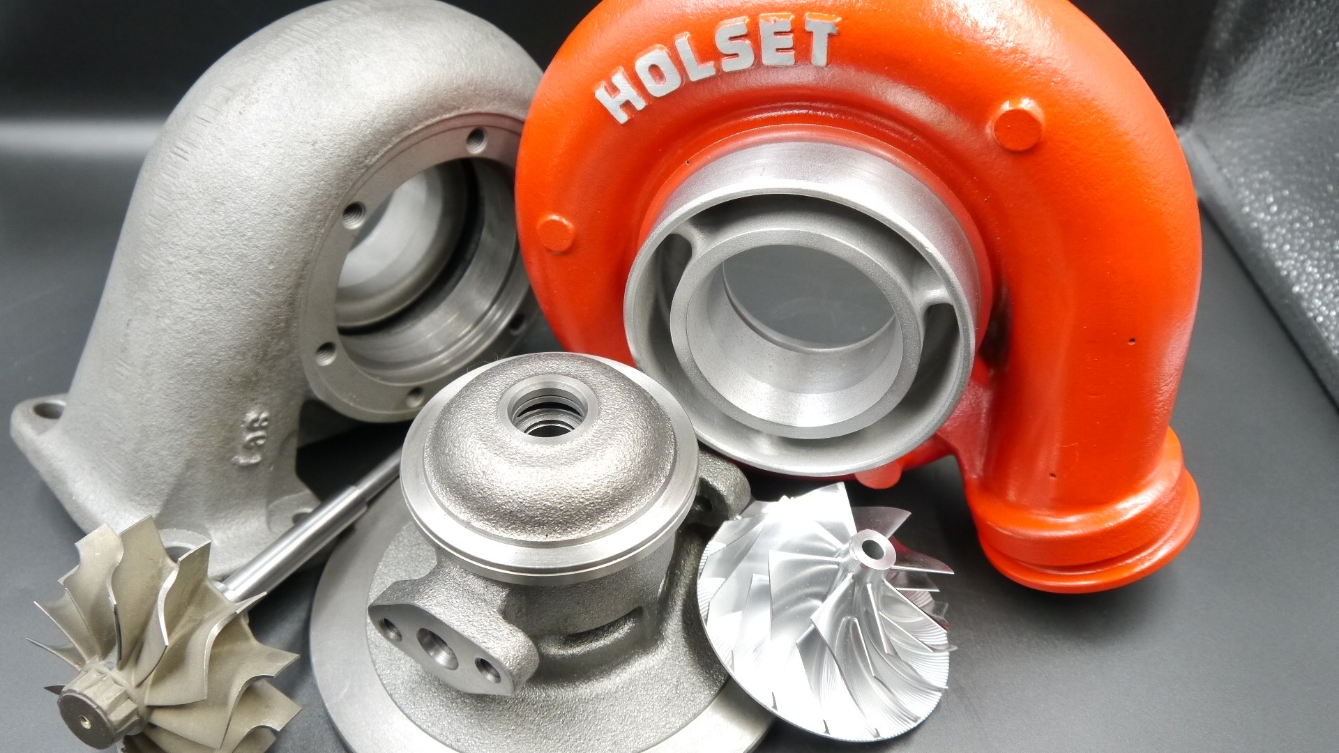 Części turbosprężarki Holset: pomarańczowa obudowa turbiny, szara obudowa kompresora oraz wirniki na ciemnym tle.