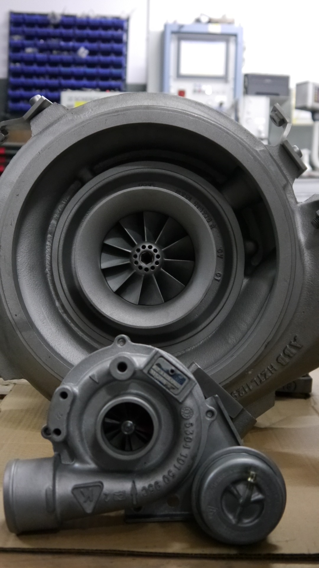 Dwa turbosprężarki: duża i mała, ustawione obok siebie na kartonowym podłożu, w tle regał z pojemnikami i urządzenie z ekranem.