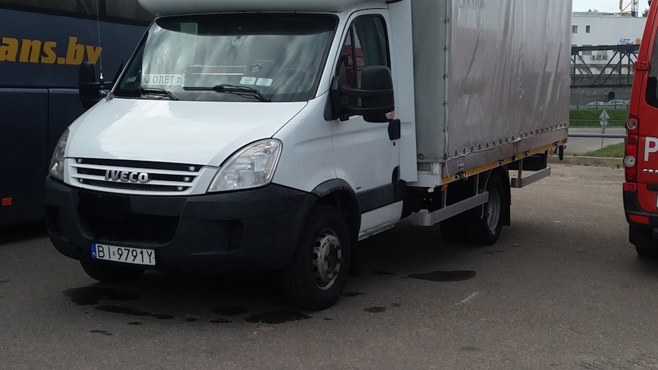 Biały samochód dostawczy Iveco z plandeką na parkingu, widoczny autobus z napisem 'trans.by' w tle.
