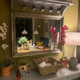 Infra-ogrzewanie - Ustawiony na szarej komodzie koszyk wiklinowy z zawartością, obok storczyki w doniczkach, flakoniki perfum i lampa z abażurem, w tle lustro odbijające jadalnię z laptopem na stole.