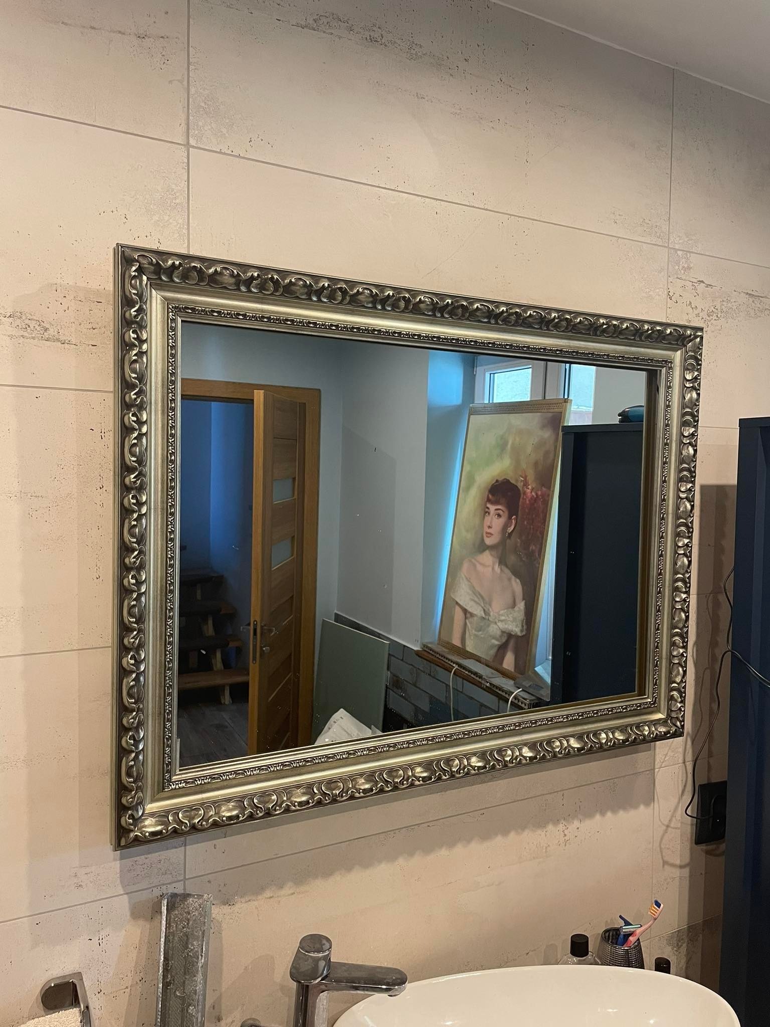 Prostokątne lustro w ozdobnej, srebrnej ramie zawieszone na ścianie wyłożonej jasnymi, kwadratowymi płytkami. W odbiciu widoczny obraz z portretem kobiety, czarny regał i otwarte drzwi prowadzące...