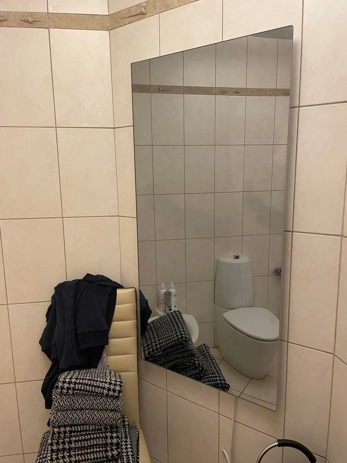 Prostokątne lustro w łazience z beżowymi płytkami, odbijające toaletę i umywalkę, z widoczną stertą ubrań na krześle po lewej stronie.