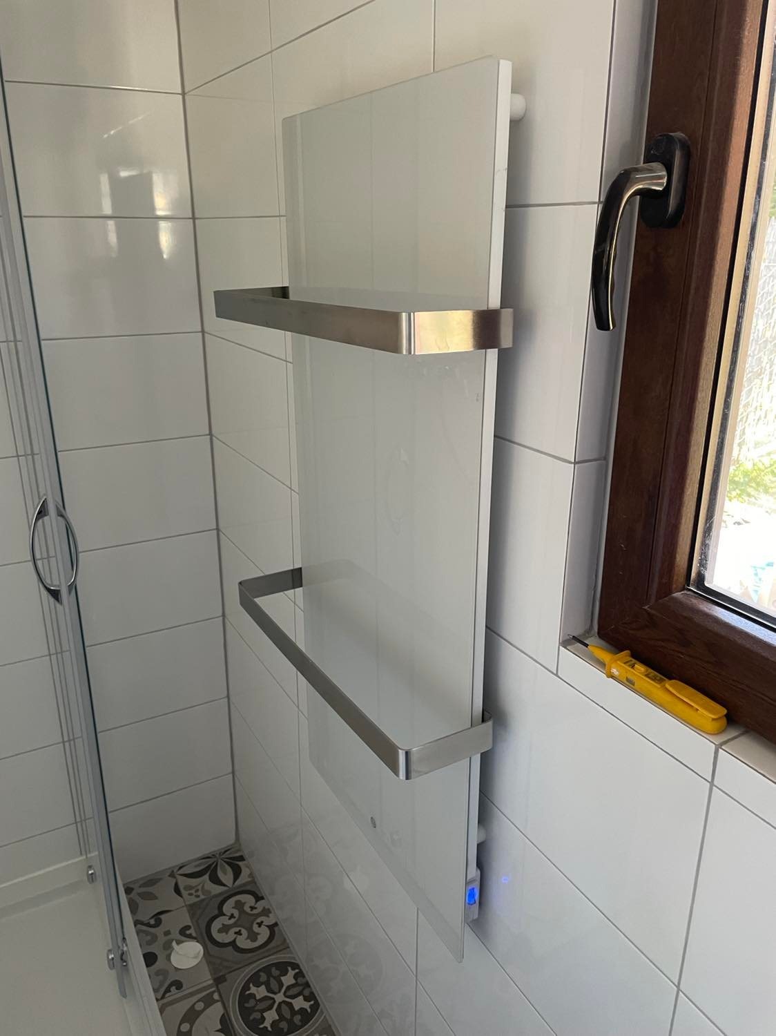 Biały, szklany grzejnik łazienkowy z dwoma chromowanymi wieszakami na ręczniki, zamontowany na ścianie z białych płytek, obok okna z brązową ramą i żółtym próbnikiem napięcia na parapecie, widoczna...