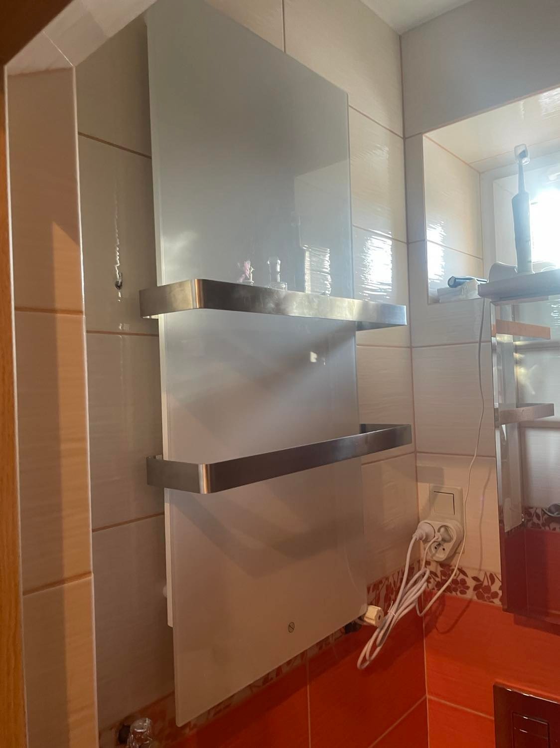 Szklany grzejnik elektryczny z dwoma chromowanymi uchwytami na ręczniki, zamontowany na kremowych płytkach w łazience, widoczne gniazdko z wtyczkami.