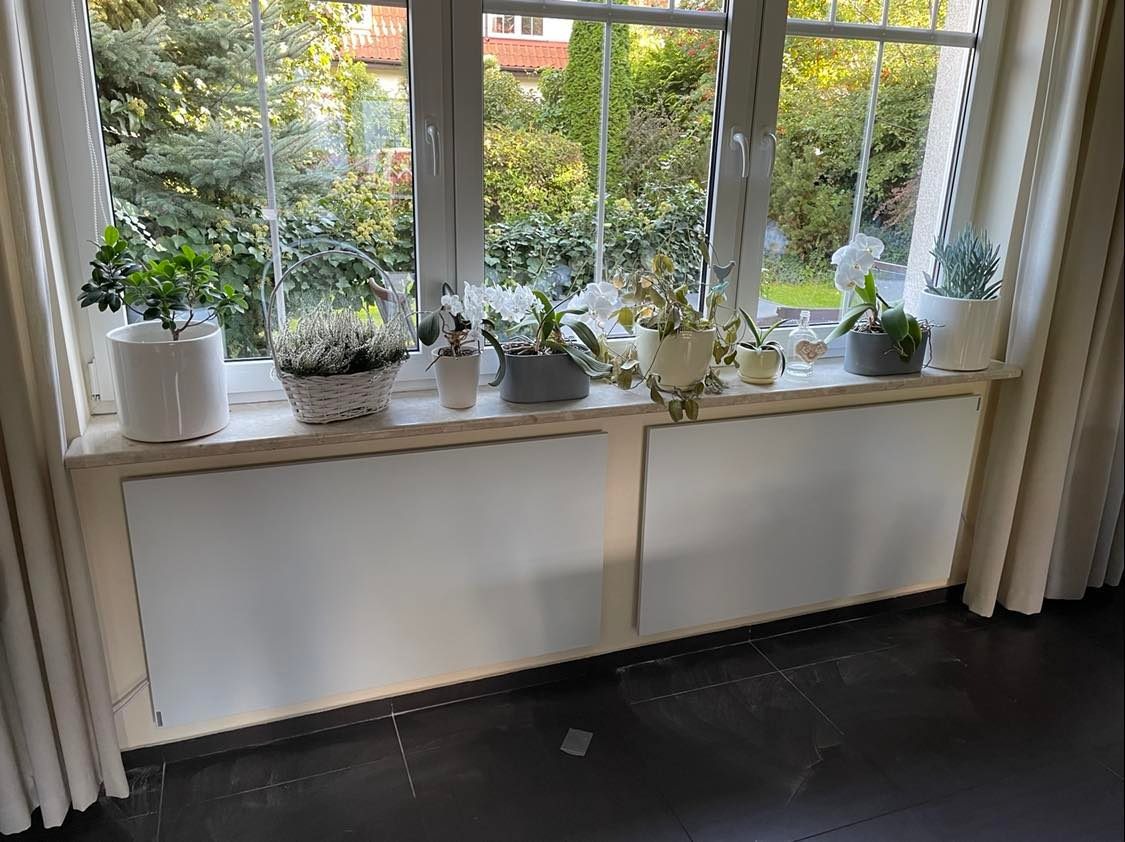 Dwa białe, płaskie panele grzewcze zamontowane pod oknem, z licznymi roślinami doniczkowymi na parapecie, widok na zielony ogród za oknem.
