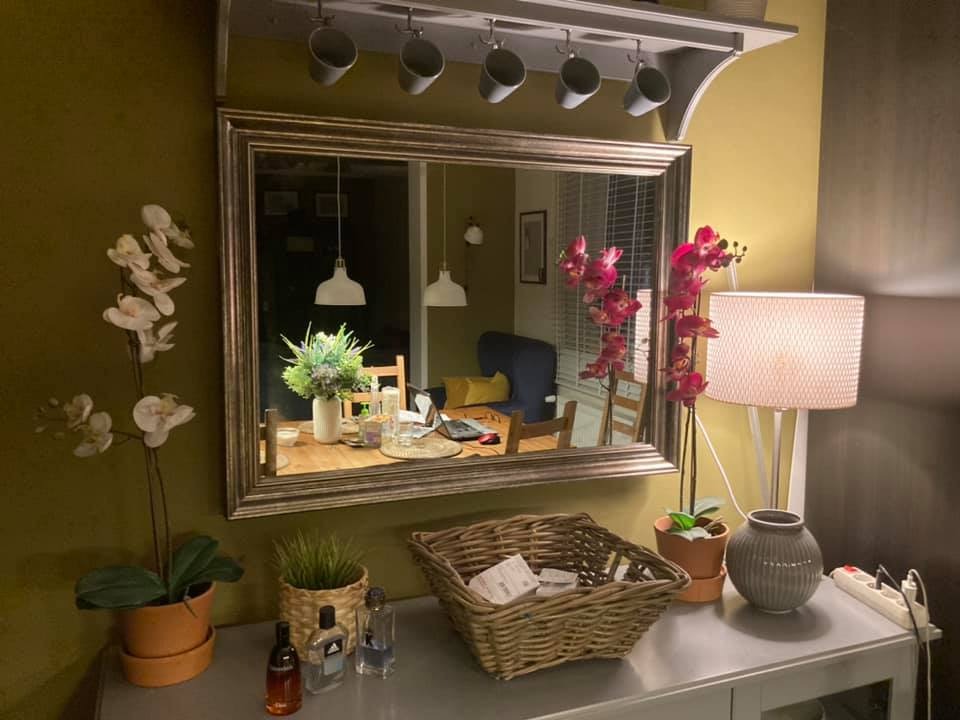 Ustawiony na szarej komodzie koszyk wiklinowy z zawartością, obok storczyki w doniczkach, flakoniki perfum i lampa z abażurem, w tle lustro odbijające jadalnię z laptopem na stole.