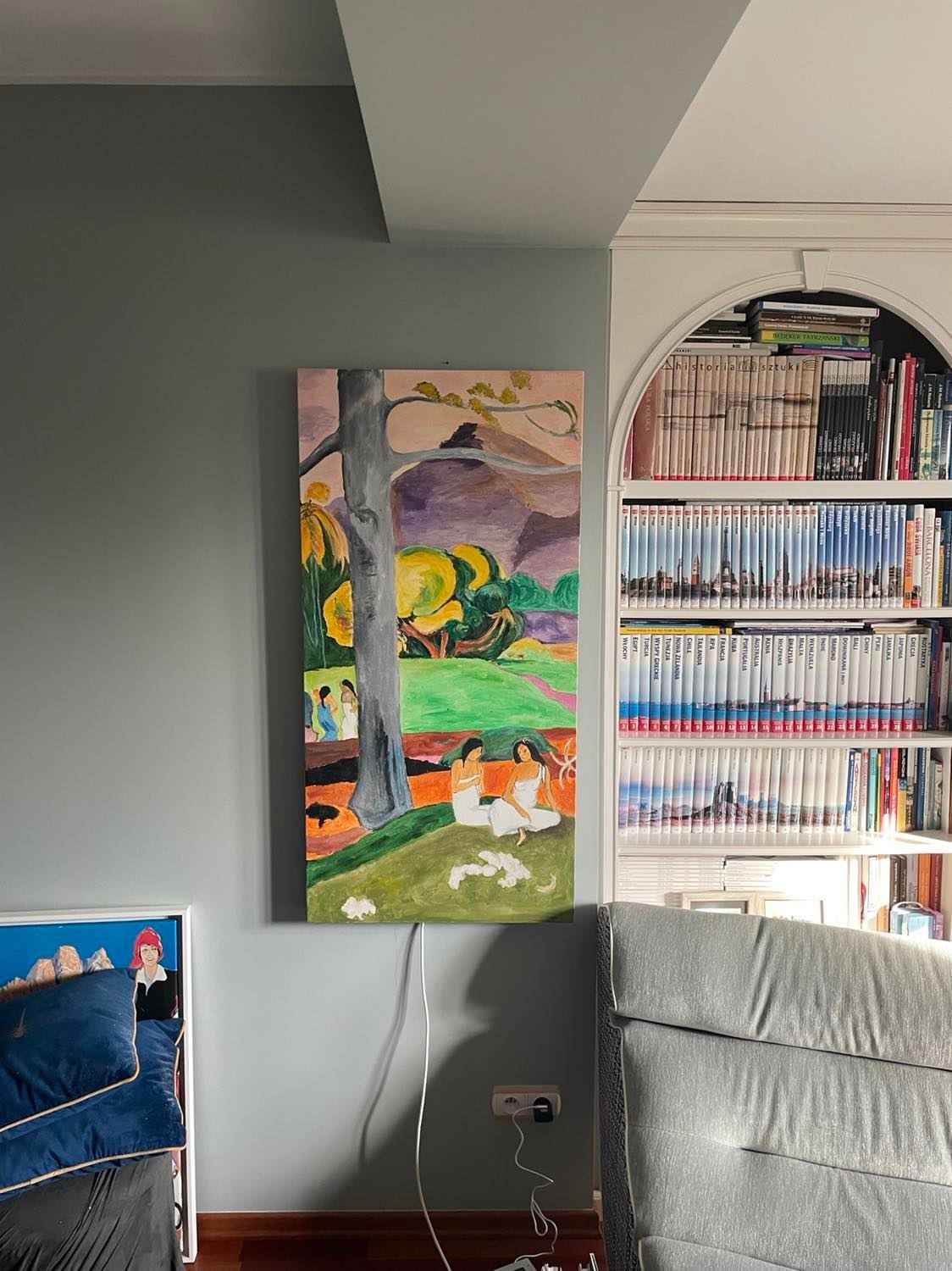 Obraz na płótnie wiszący na szarej ścianie, przedstawiający scenę z drzewem, postaciami i krajobrazem w stylu Gauguina, z widocznymi kablami zasilającymi i fragmentem regału z książkami w tle.