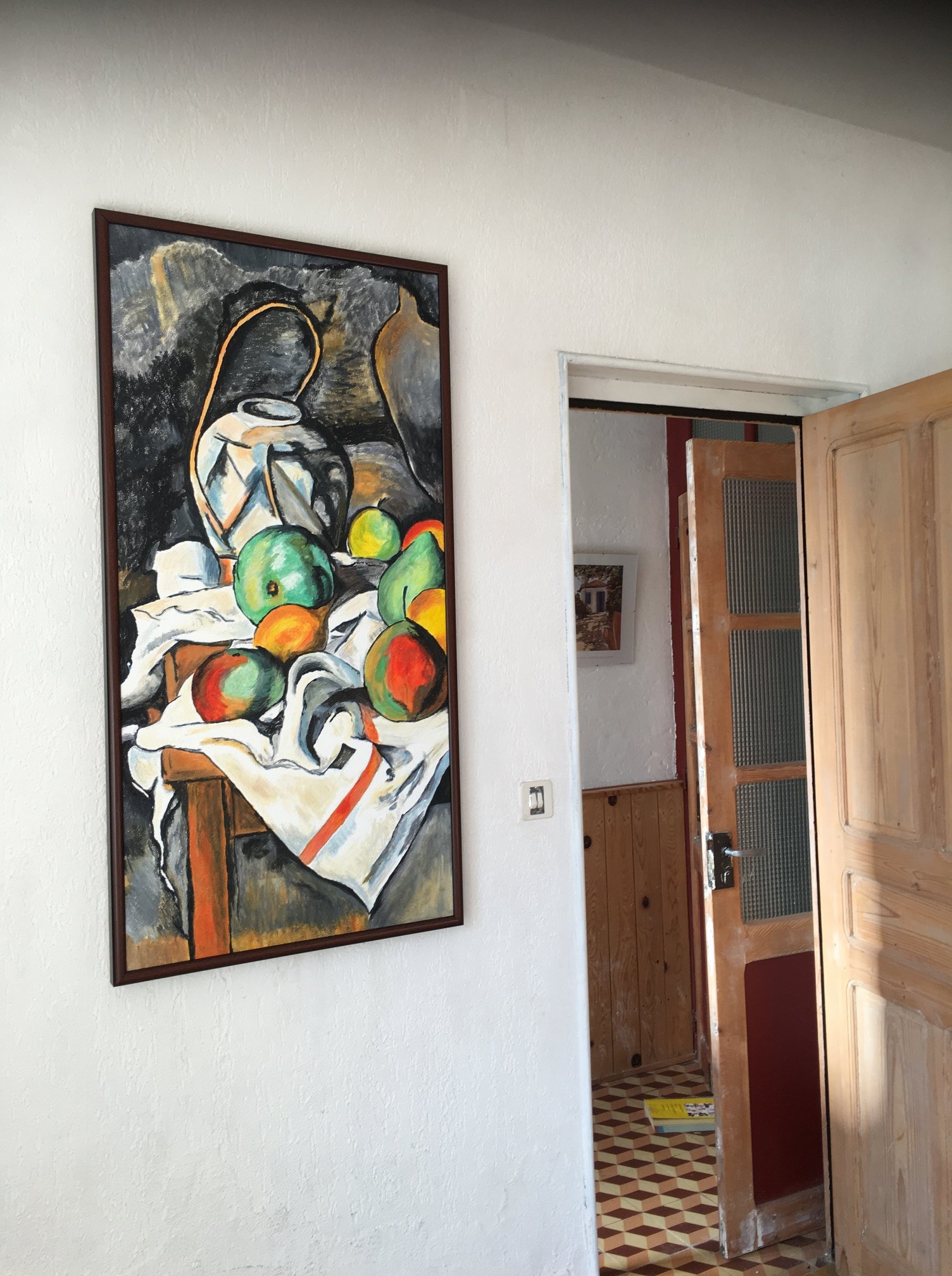 Obraz w brązowej ramie przedstawiający martwą naturę z dzbankiem i owocami, wiszący na białej ścianie nad wejściem do pokoju z drewnianą podłogą w kratkę i drewnianą boazerią.