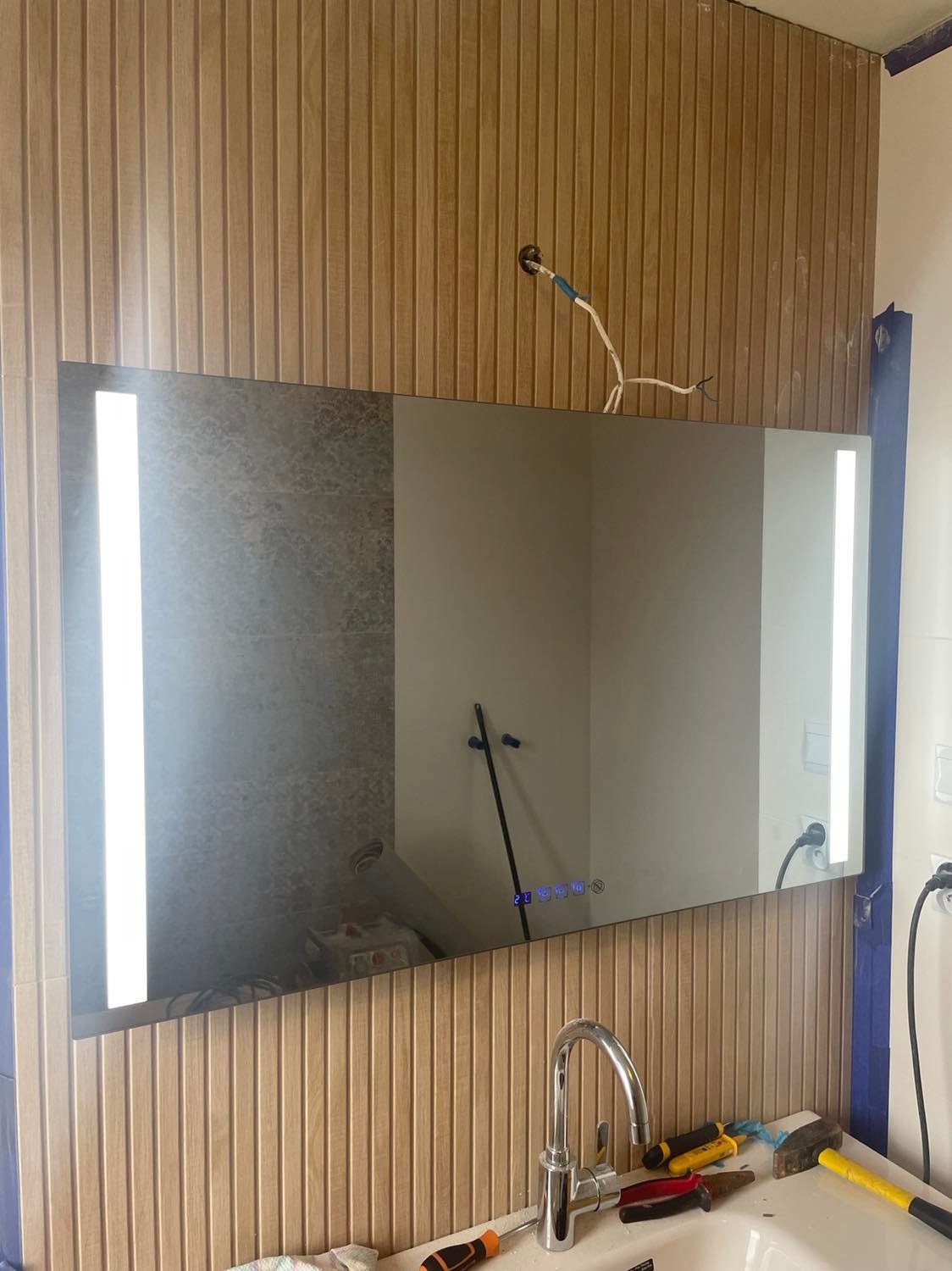 Montaż podświetlanego lustra łazienkowego na ścianie z drewnianymi panelami, widoczny kran, narzędzia i niedokończone podłączenie elektryczne.