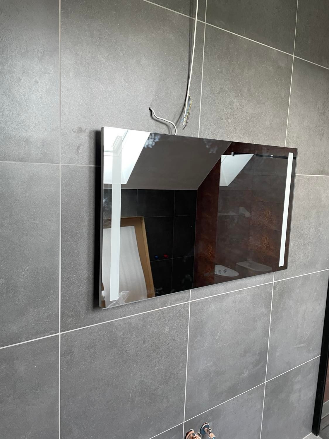 Prostokątne lustro z podświetleniem LED zamontowane na szarej, kafelkowanej ścianie, widoczne kable instalacyjne, odbicie wnętrza łazienki z elementami wyposażenia.