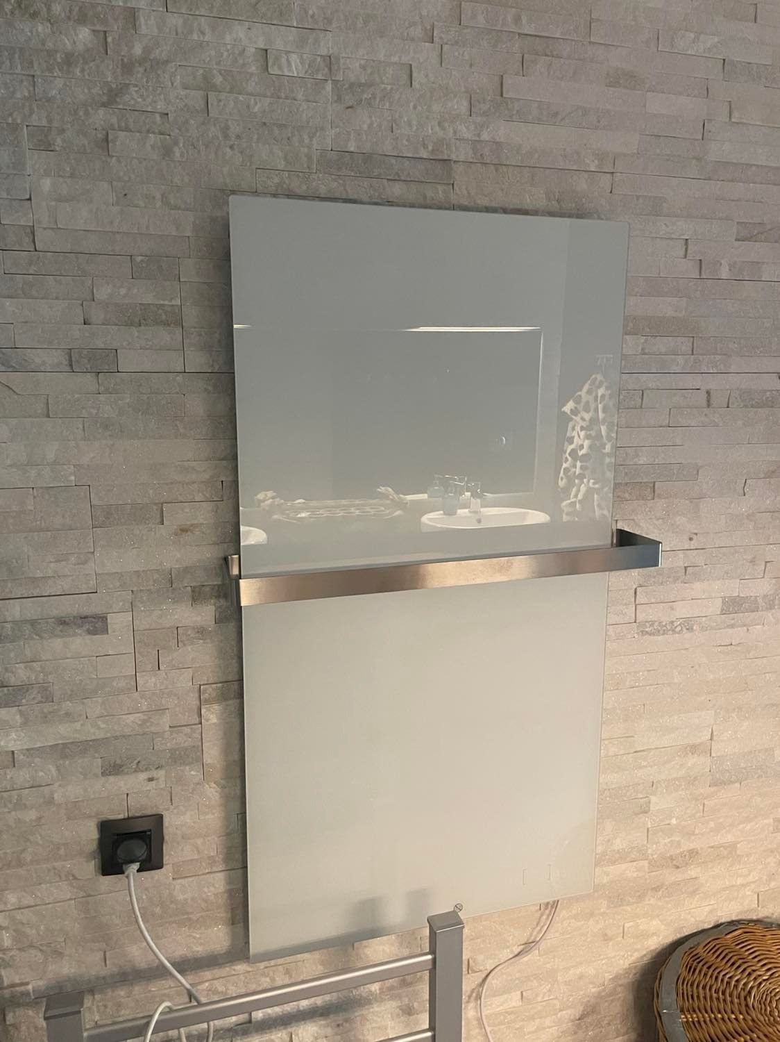 Szklany grzejnik panelowy z metalowym uchwytem na ręczniki, zamontowany na ścianie z dekoracyjnej cegły, widoczny kabel zasilający i fragment wieszaka na ręczniki.