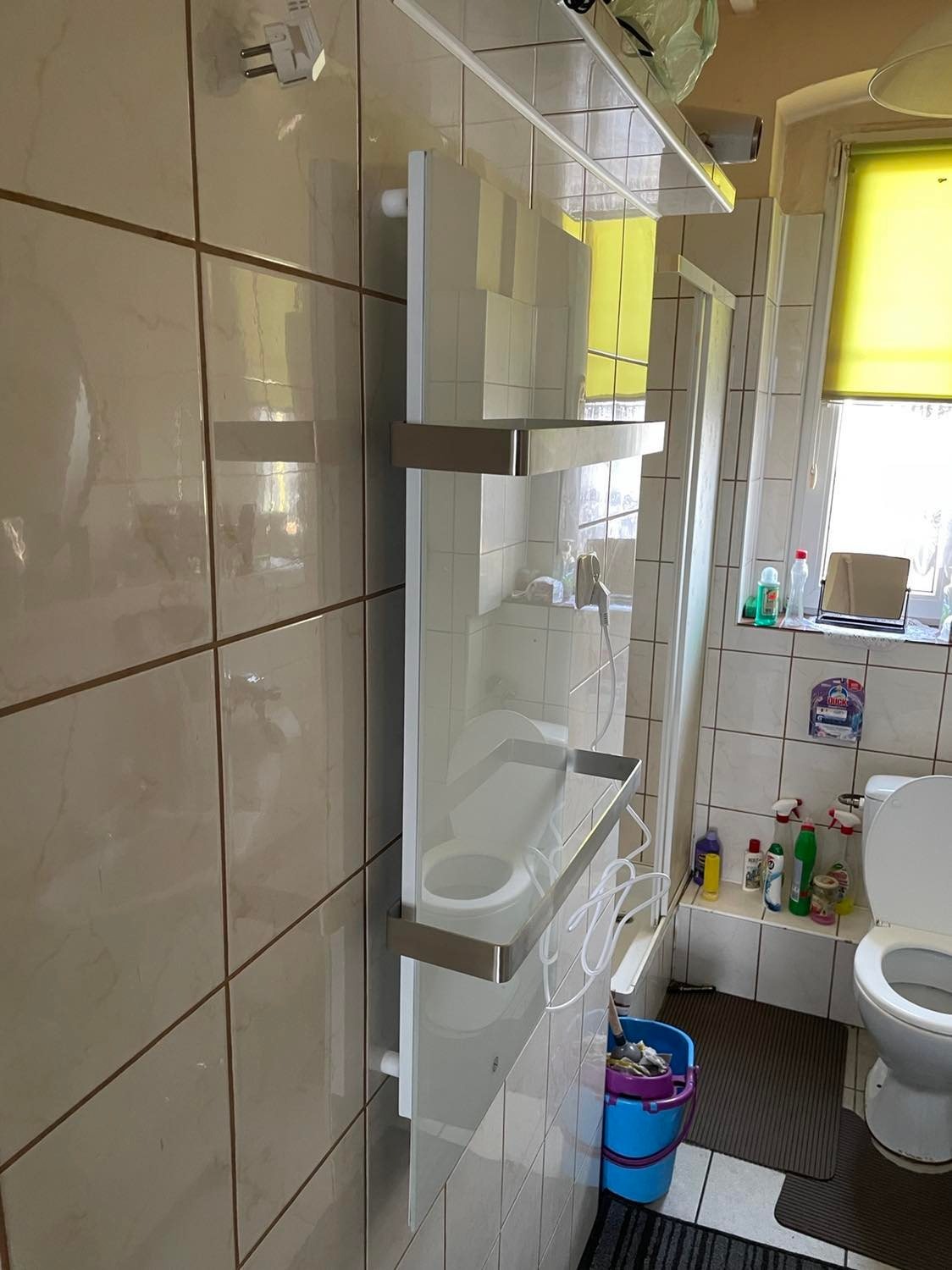 Biały, nowoczesny grzejnik łazienkowy z metalowymi wstawkami zamontowany na ścianie z beżowych płytek w łazience z widoczną toaletą, kabiną prysznicową i oknem z żółtą roletą.