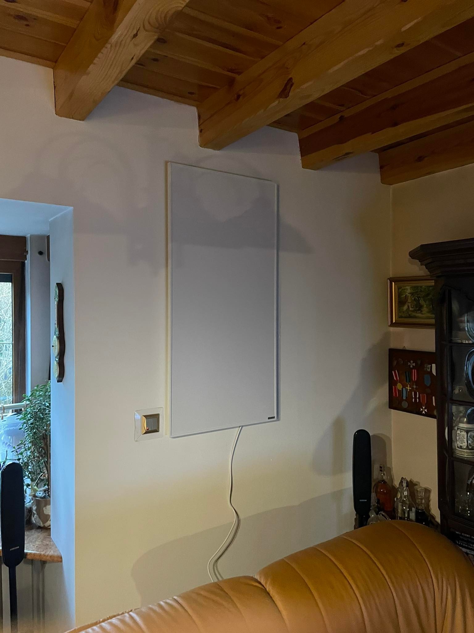 Biały panel grzewczy na ścianie w jasnym wnętrzu z drewnianym stropem, widoczny kabel zasilający i włącznik światła obok.