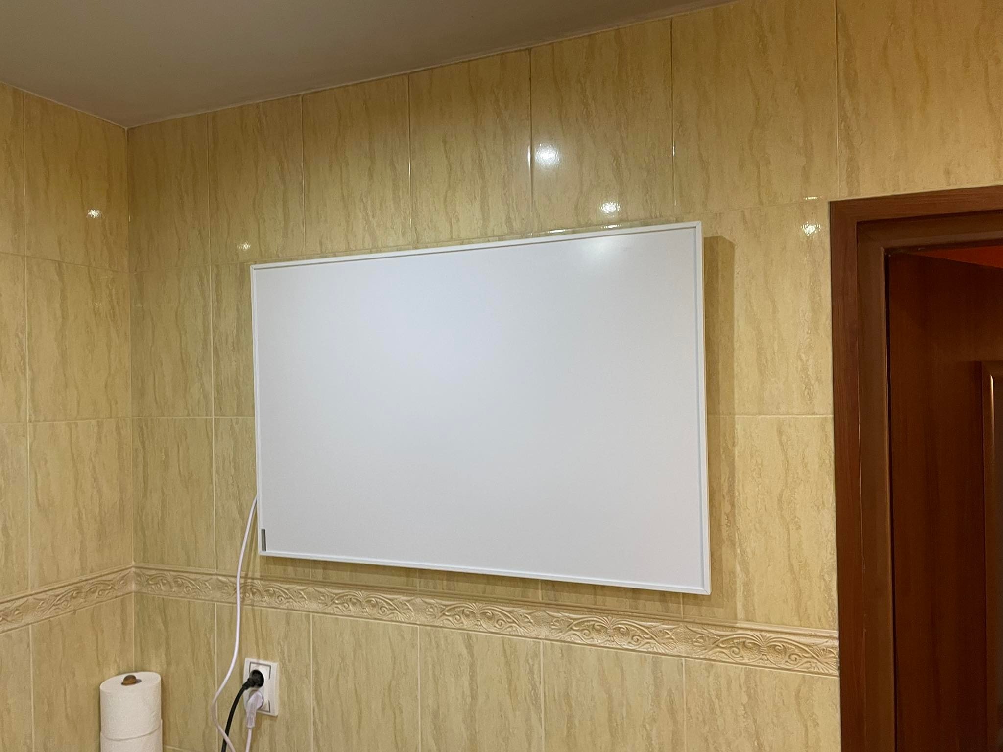 Płaski, biały panel grzewczy elektryczny zamontowany na ścianie wyłożonej beżowymi, marmurkowymi płytkami ceramicznymi w łazience, widoczny kabel zasilający i gniazdko elektryczne.
