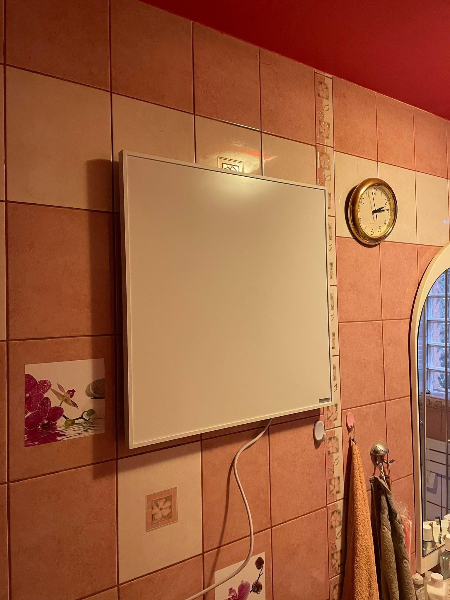 Płaski, biały panel grzewczy zawieszony na ścianie z brązowych płytek w łazience, widoczny zegar i fragment lustra.
