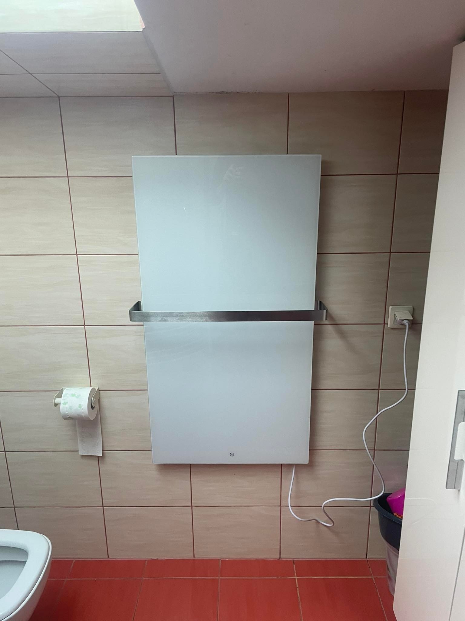 Biały, elektryczny grzejnik łazienkowy z chromowanym wieszakiem na ręczniki, zamontowany na kafelkowanej ścianie w łazience z czerwonymi płytkami podłogowymi.