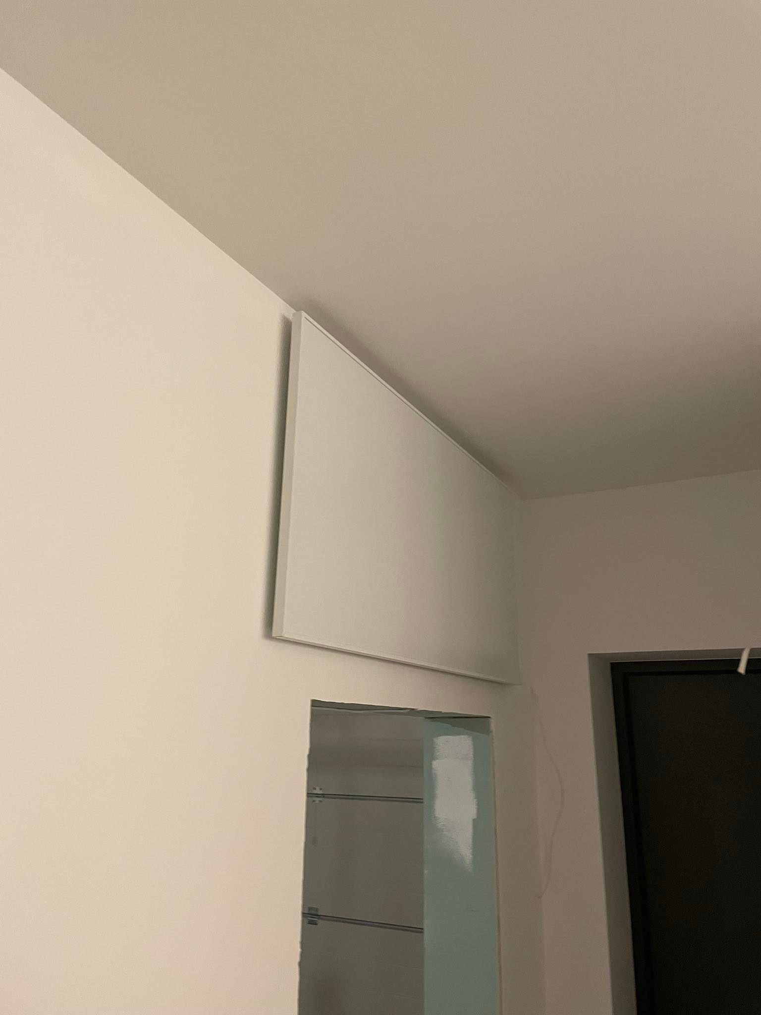 Płaski, biały grzejnik panelowy zamontowany na ścianie nad otworem w ścianie z widoczną konstrukcją wewnątrz, obok czarnej futryny drzwiowej.