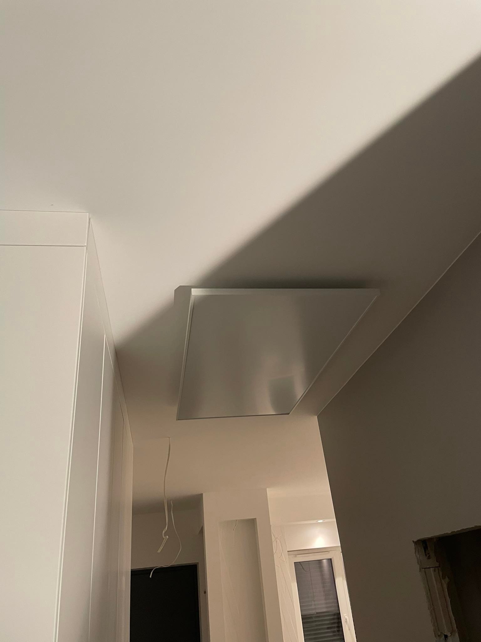 Płaski, prostokątny panel grzewczy zamontowany na suficie, widoczne wiszące kable, fragment zabudowy meblowej w kolorze białym i ściana w odcieniach szarości.