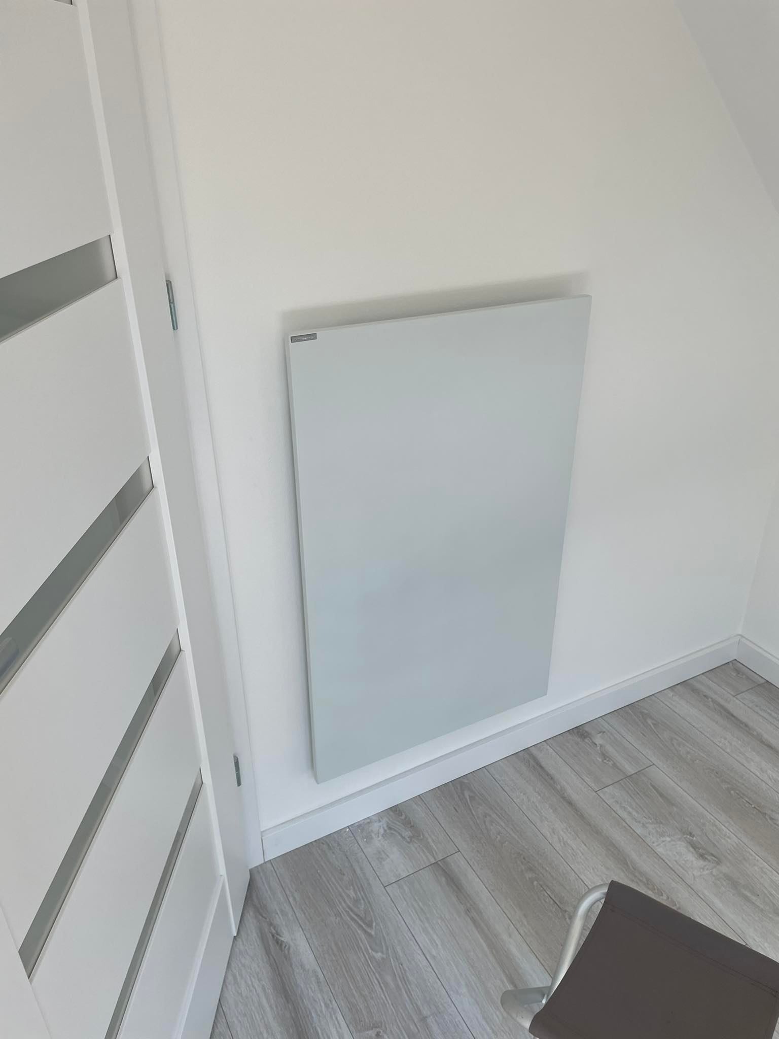 Płaski, panelowy grzejnik elektryczny w jasnym pomieszczeniu, widoczny fragment białych drzwi z poziomymi wstawkami i szare panele podłogowe.