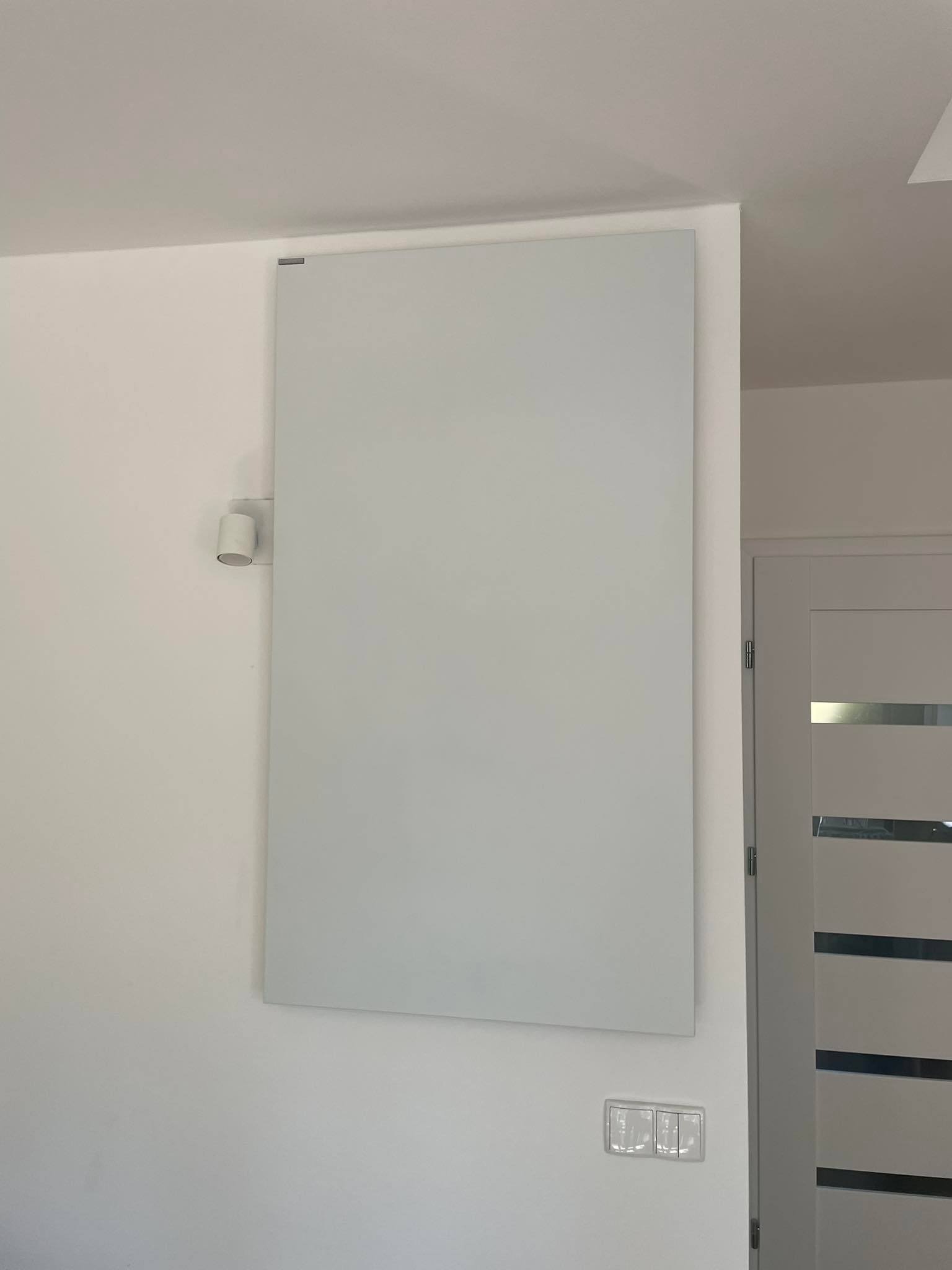 Biały, płaski grzejnik elektryczny zamontowany na ścianie w nowoczesnym wnętrzu, widoczny włącznik światła i fragment białych drzwi z poziomymi, szarymi paskami.