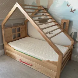 Łóżko dziecięce w kształcie domku z jasnego drewna, z drabinką wejściową i małym okienkiem, na tle ściany z motywem balonu i ptaków.