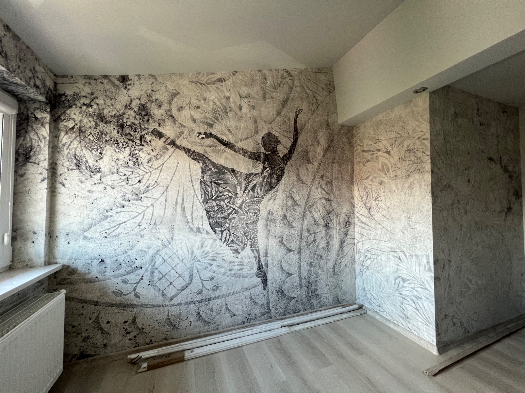 Pokój po remoncie z tapetą przedstawiającą sylwetkę baletnicy na tle roślinnych wzorów, ułożone rolki tapety przy ścianie, grzejnik pod oknem.