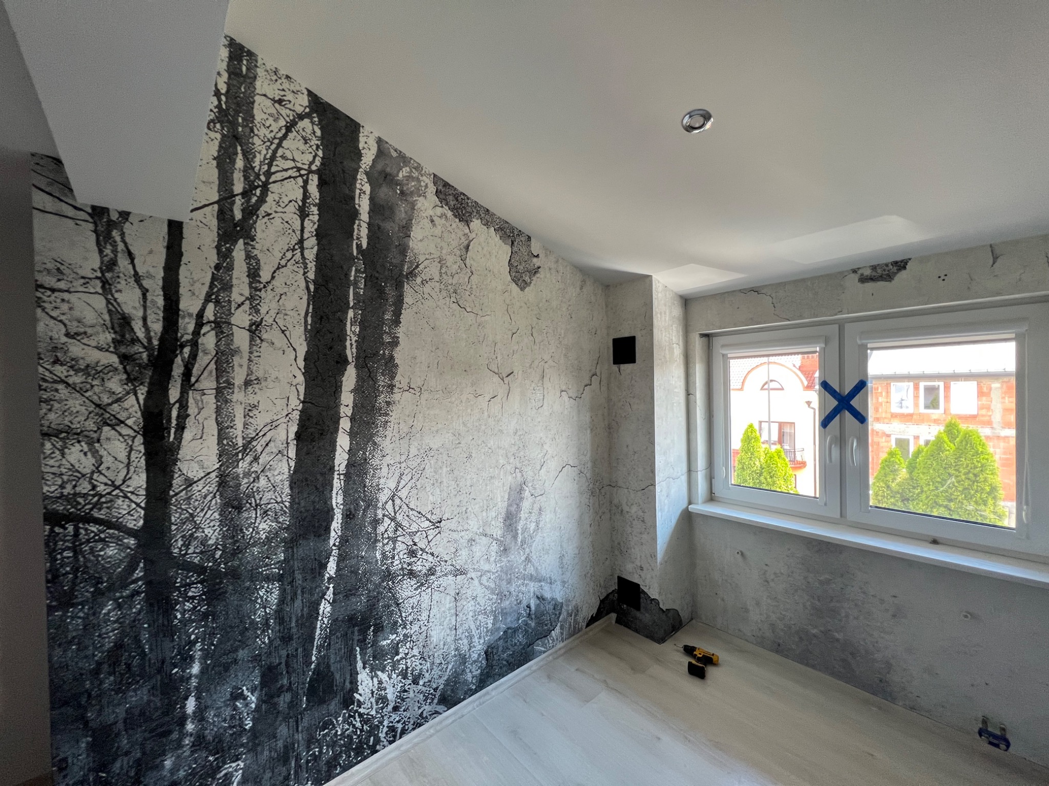 Pokój w trakcie remontu, ściana pokryta fototapetą z motywem lasu, widoczne narzędzia i niedokończone prace wykończeniowe przy oknie.