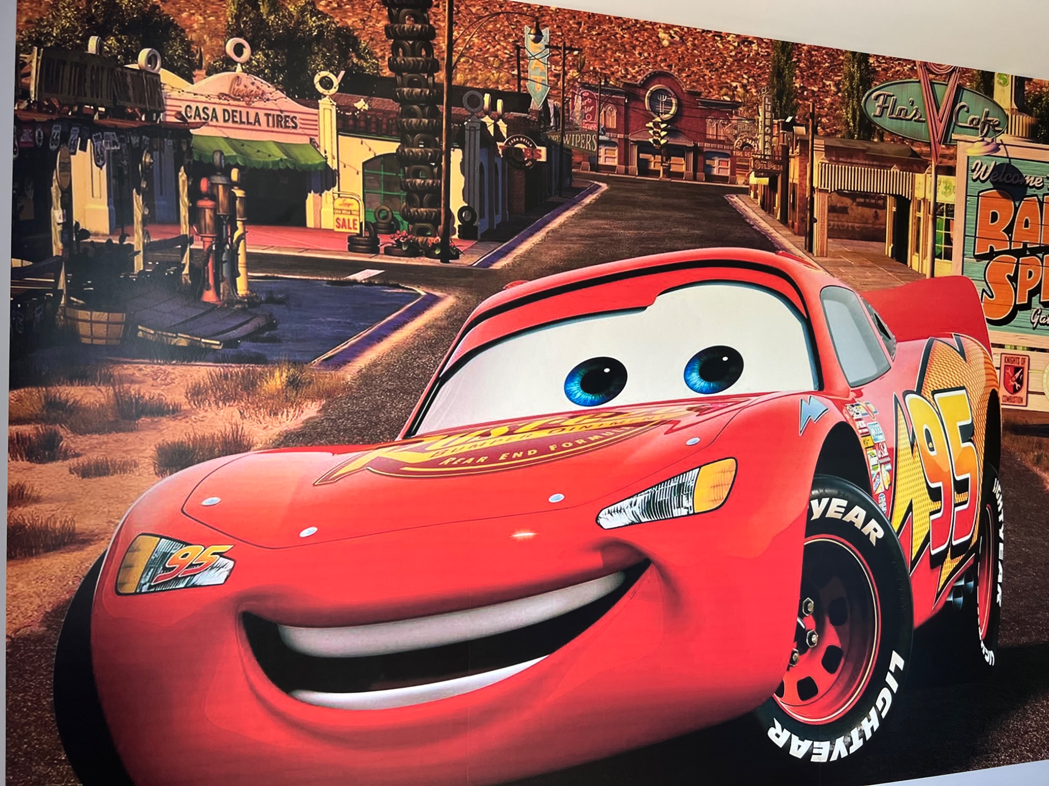 Zdjęcie przedstawia fototapetę z motywem z filmu Auta, na której widoczny jest Zygzak McQueen na tle miasteczka Chłodnica Górska z Casa Della Tires i Flos Cafe.