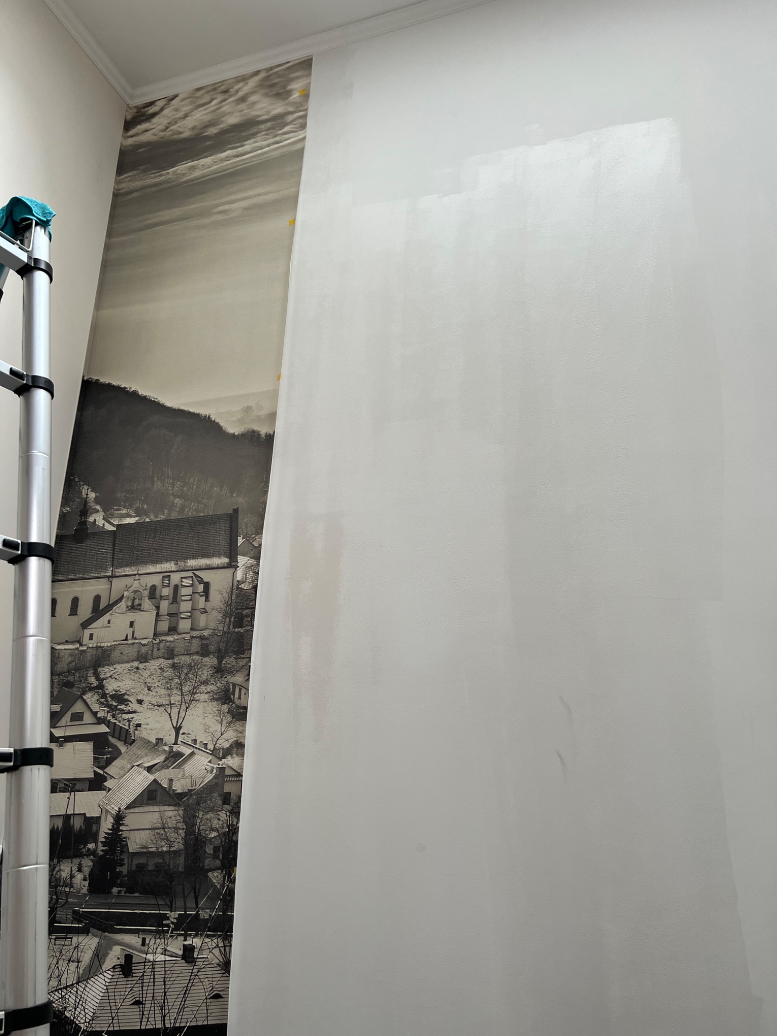 Fragment ściany z częściowo nałożoną fototapetą przedstawiającą czarno-biały krajobraz z budynkami i wzgórzami, widoczna drabina aluminiowa z niebieską szmatką na górze.