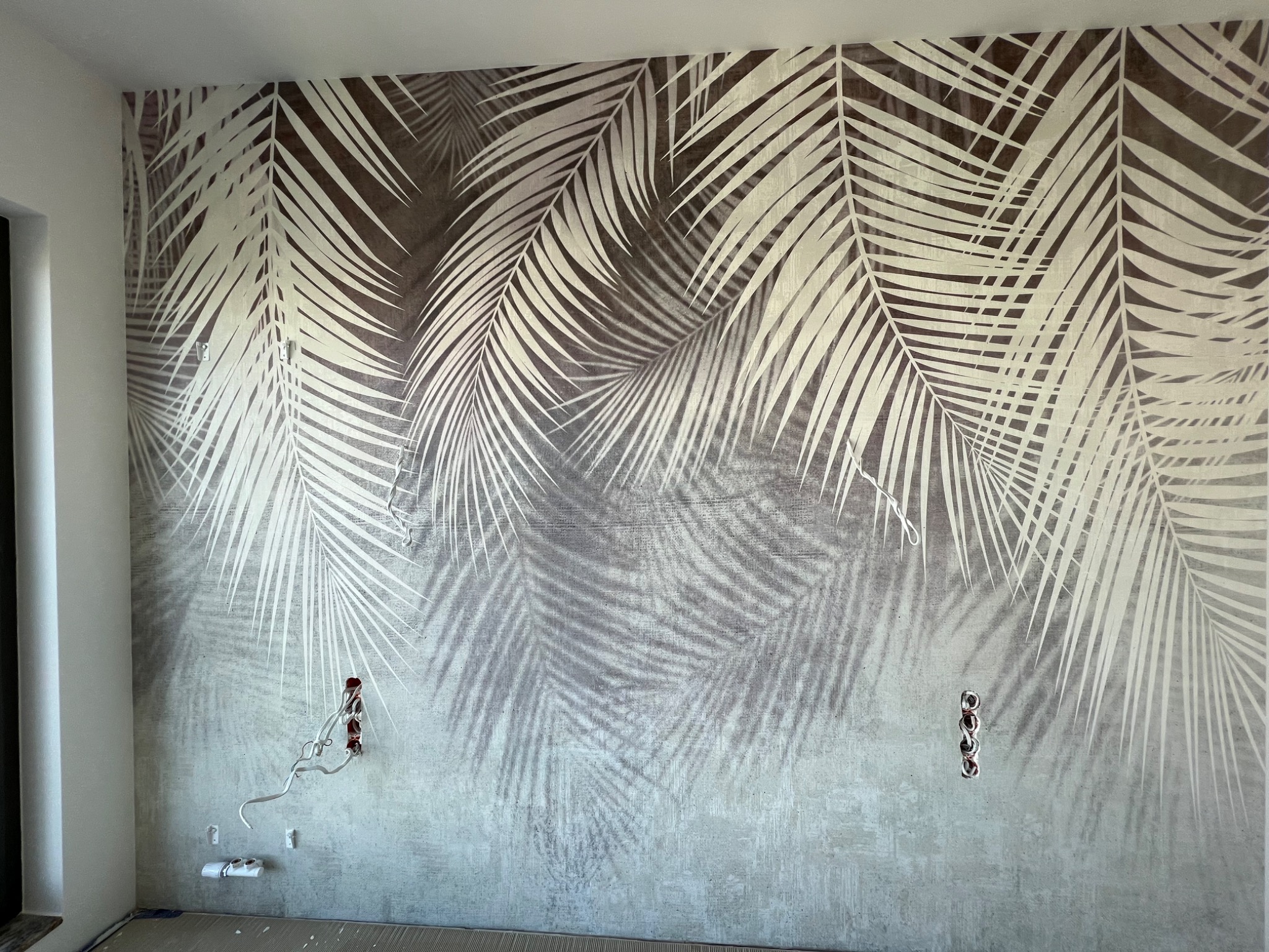 Ściana po nałożeniu tapety z motywem liści palmowych w odcieniach szarości i bieli, widoczne wystające kable elektryczne.