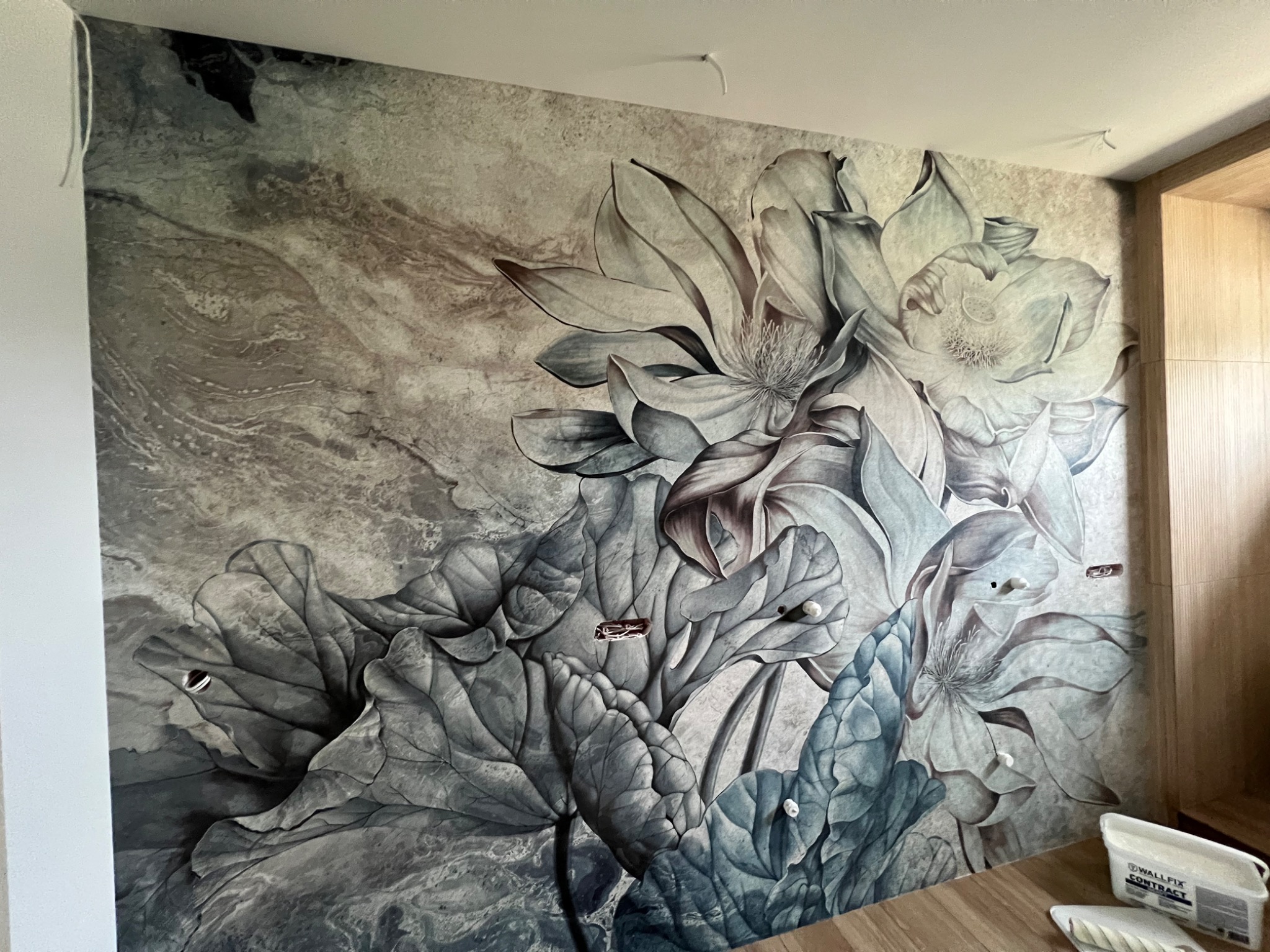 Ściana pokryta fototapetą z motywem kwiatowym w odcieniach szarości i błękitu, widoczne niedoskonałości w postaci otworów na gniazdka elektryczne i akcesoria do montażu na drewnianej podłodze.