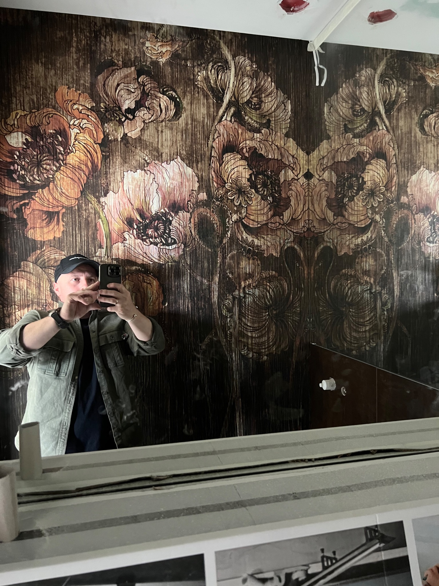 Mężczyzna w czapce i zielonej kurtce robi sobie selfie na tle ściany pokrytej tapetą w ciemne kwiatowe wzory, widoczne niedociągnięcia w górnej części ściany.