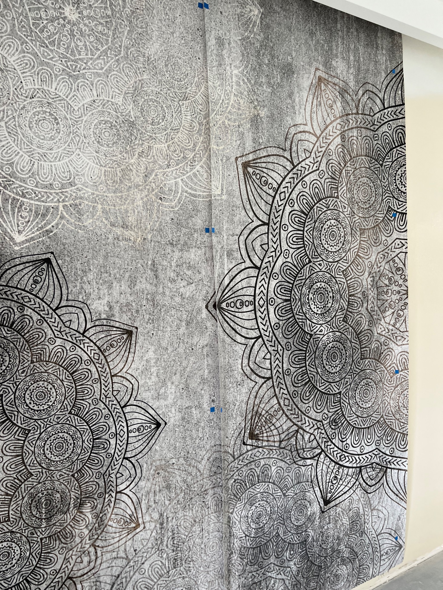Tapeta z szarym tłem imitującym beton i dużymi, symetrycznymi wzorami mandali w kolorze czarnym i białym, widoczne niebieskie znaczniki montażowe, fragment ściany pomalowanej na kremowo po prawej...