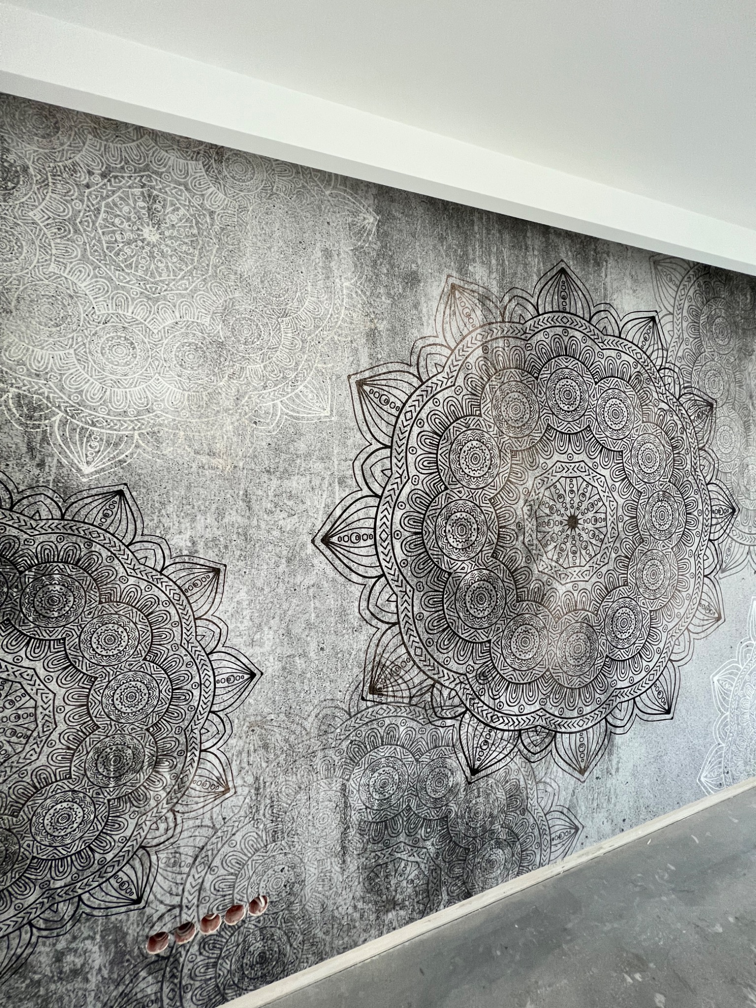 Ściana pokryta szarą tapetą z dużymi, symetrycznymi wzorami mandali w odcieniach bieli i czerni, widoczne otwory na gniazdka elektryczne w dolnej części ściany.