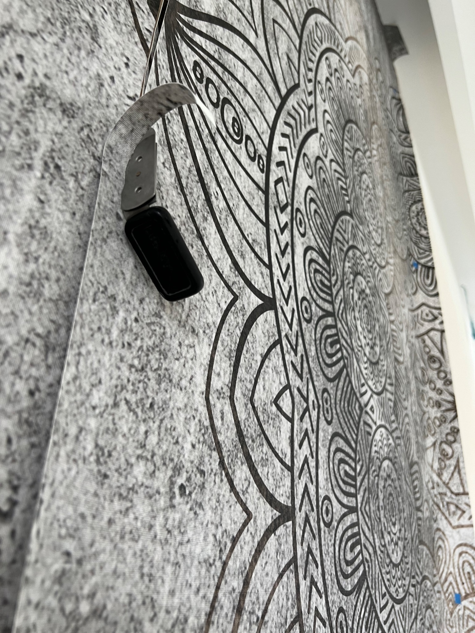 Narzędzie do tapetowania przytrzymujące fragment tapety z motywem mandali na szarej ścianie z widoczną strukturą.