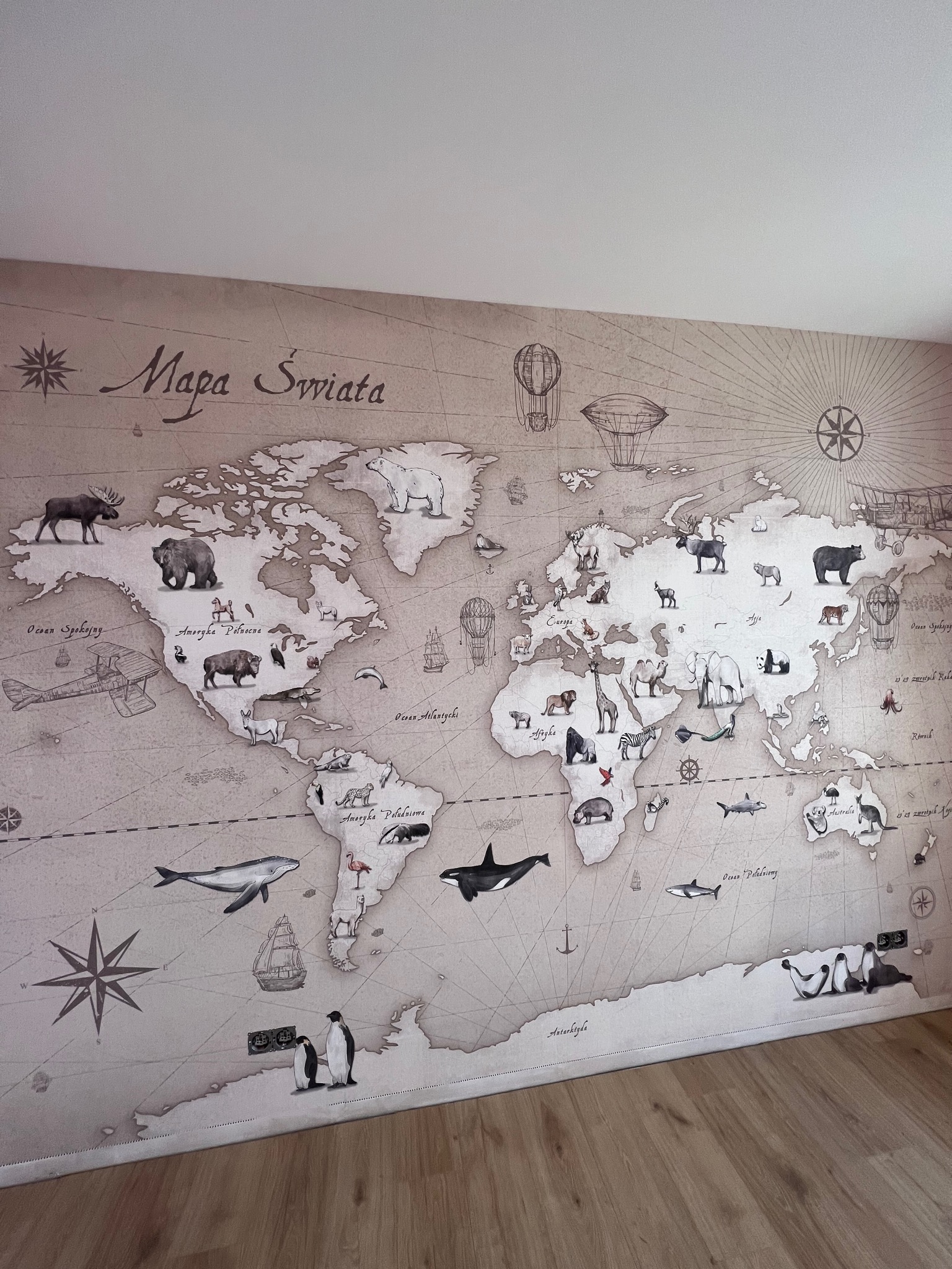 Ściana pokryta tapetą z motywem mapy świata w stylu vintage, z ilustracjami zwierząt charakterystycznych dla różnych kontynentów i konturami kontynentów. Na podłodze widoczne panele podłogowe.
