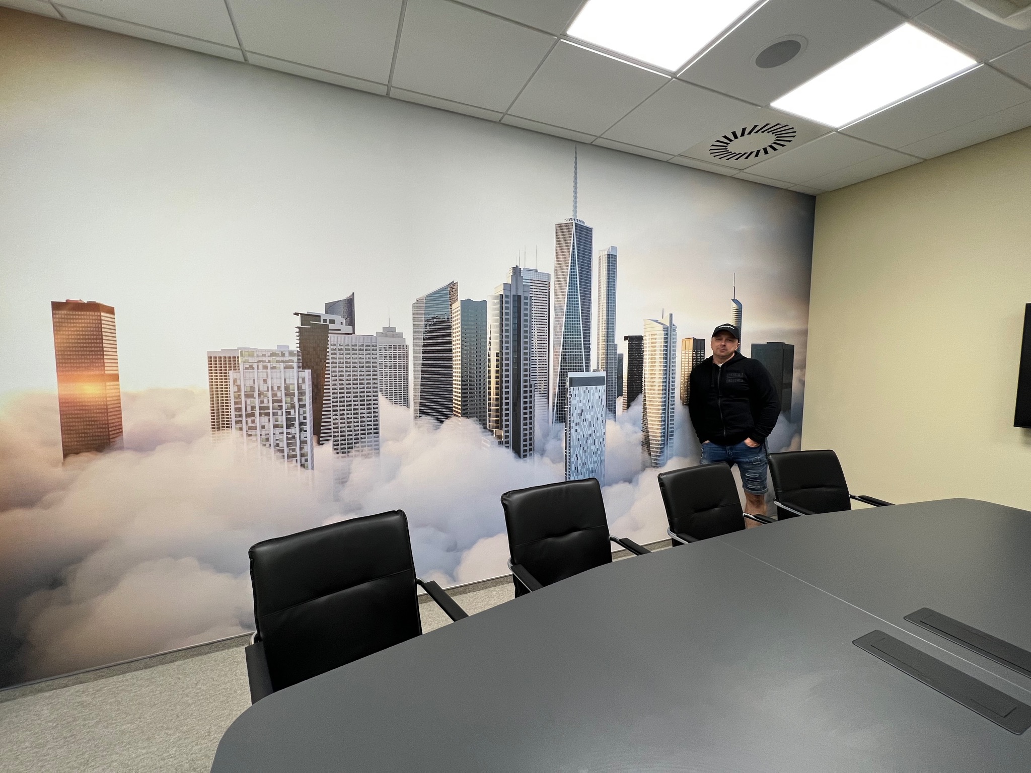 Biuro z fototapetą przedstawiającą panoramę miasta w chmurach, czarne krzesła konferencyjne i mężczyzna w czarnej bluzie stojący przy ścianie.