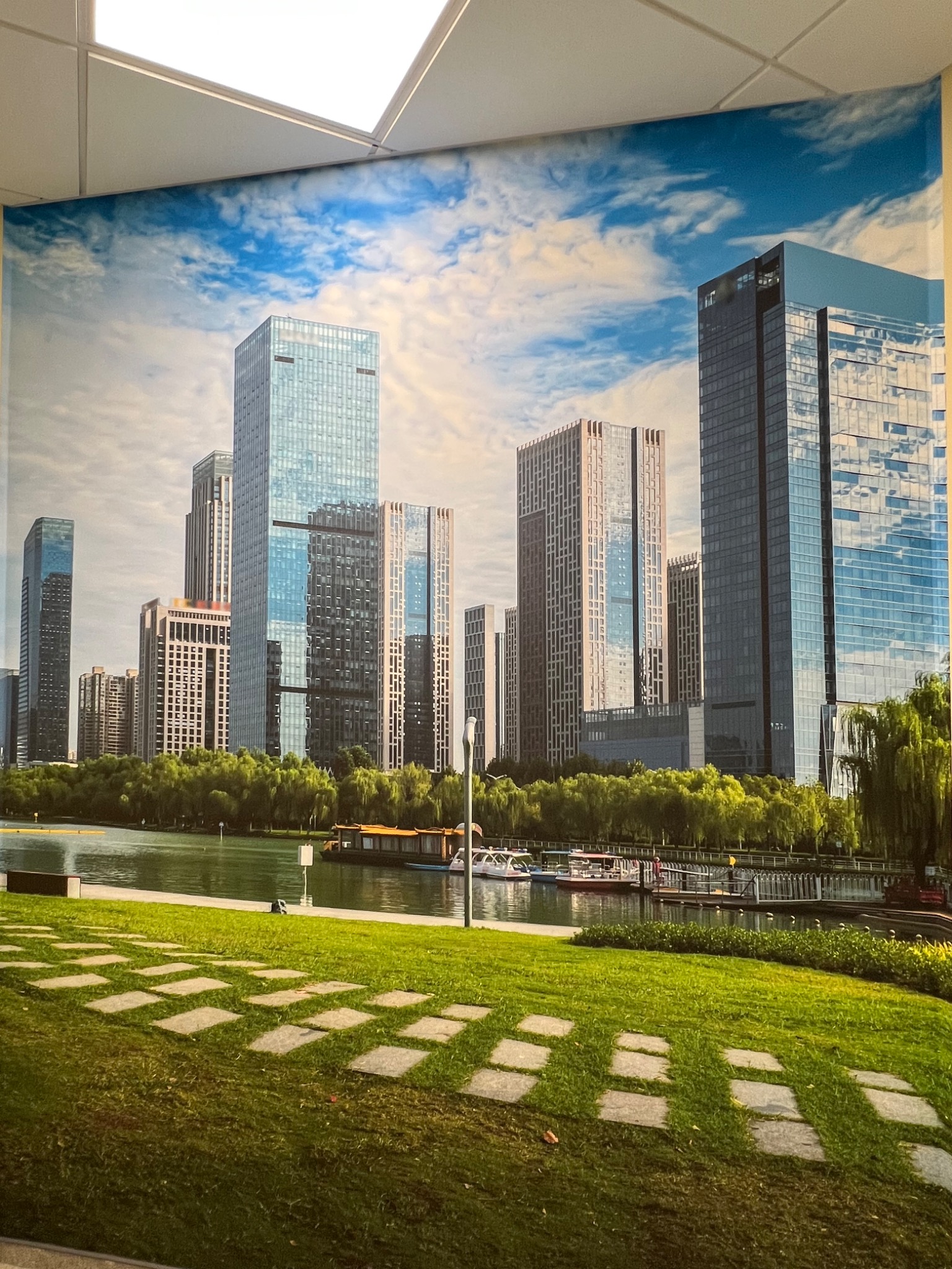 Fototapeta z motywem nowoczesnego miasta z wieżowcami, jeziorem i zielenią, zamontowana na ścianie pod sufitem.