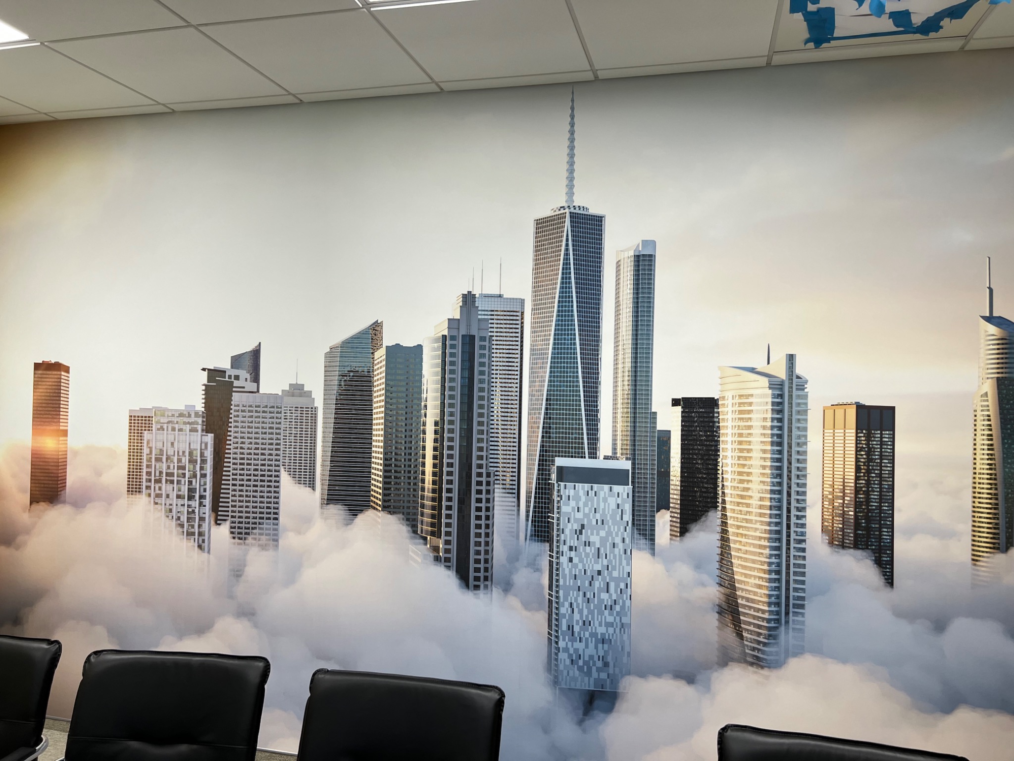 Fototapeta w biurze przedstawiająca panoramę miasta z wieżowcami wystającymi ponad chmury, widok zza czarnych krzeseł biurowych, oświetlenie sufitowe.
