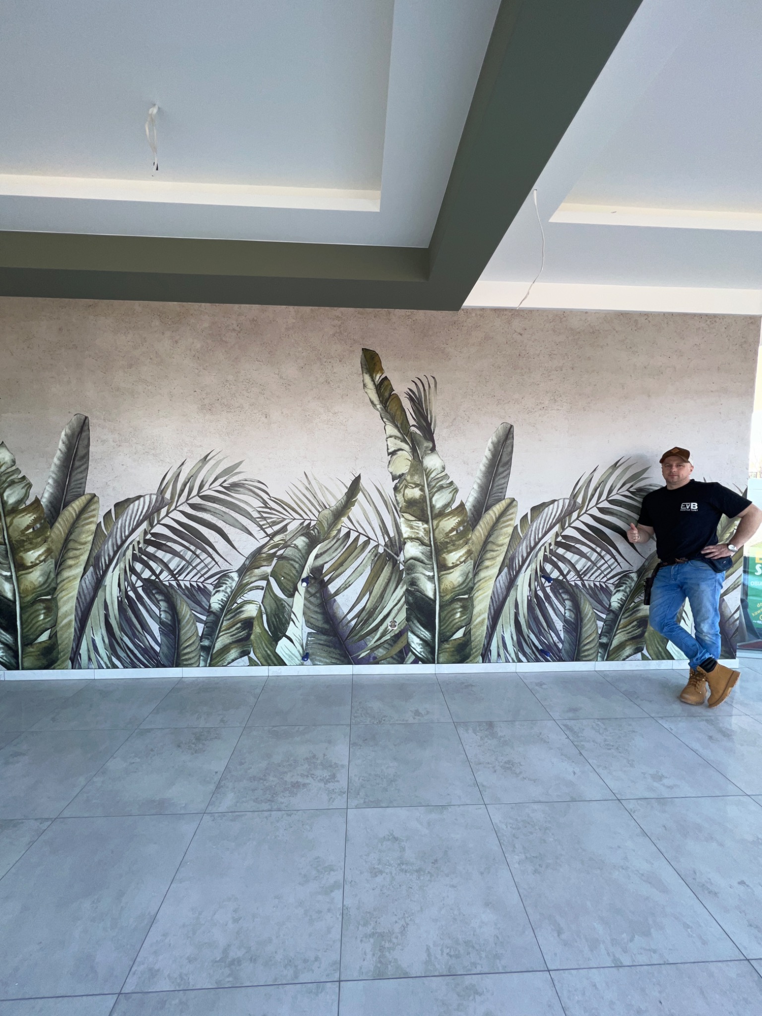 Mężczyzna oparty o ścianę z fototapetą w motywie tropikalnych liści, na podłodze szare płytki
