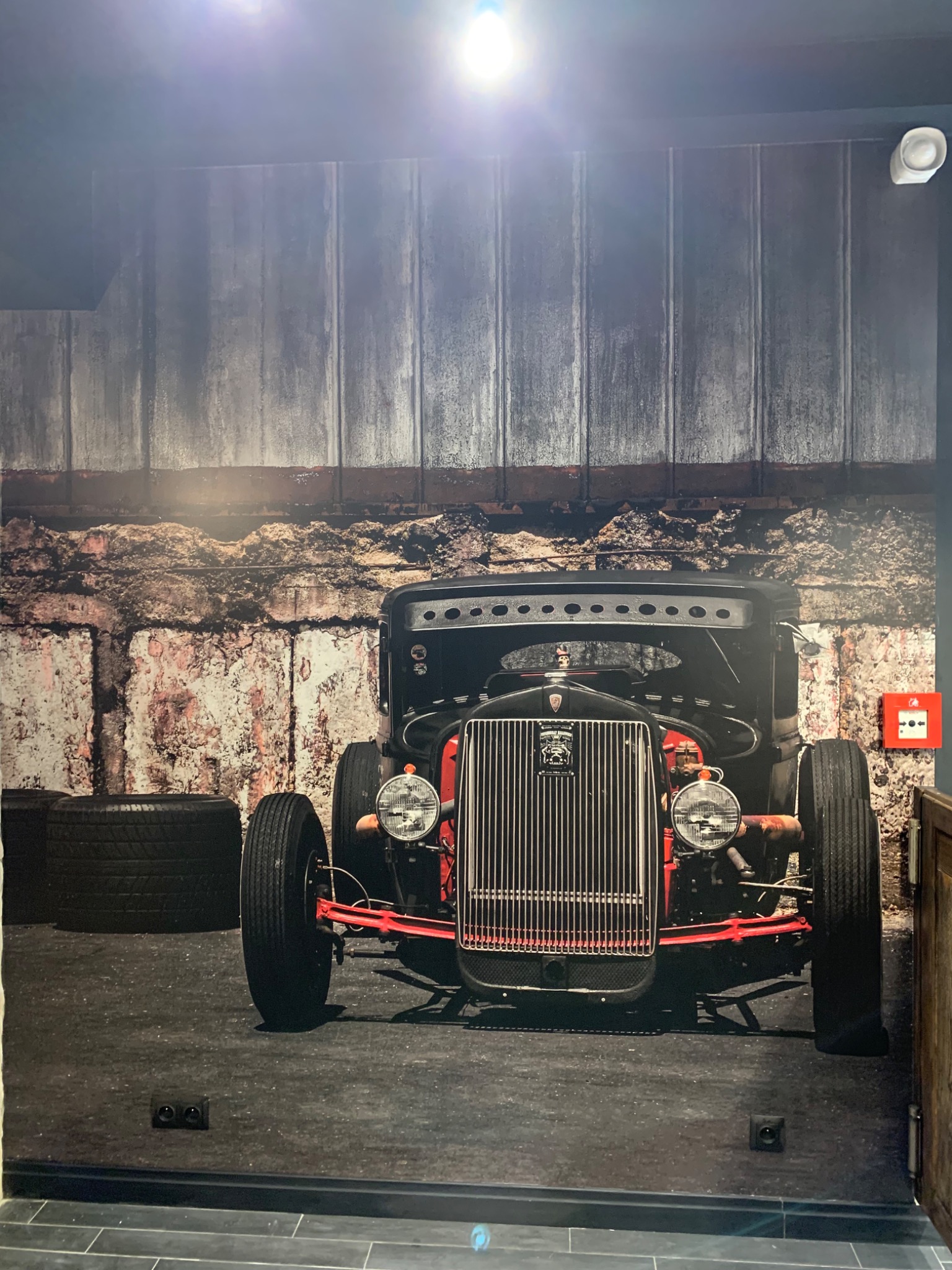 Fototapeta w Lublinie przedstawiająca czarny, klasyczny hot rod na tle imitacji starej, ceglanej ściany i drewnianych desek. Widoczne opony i czerwone elementy podwozia.