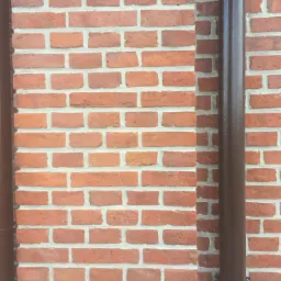 Fragment ściany z czerwonej cegły z widocznymi brązowymi rurami spustowymi po bokach.