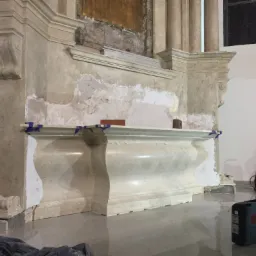 Renowacja marmurowego ołtarza w kościele, widoczne ślady napraw i uzupełnień, zabezpieczone taśmą malarską, sprzęt budowlany w tle, odblaskowa posadzka.